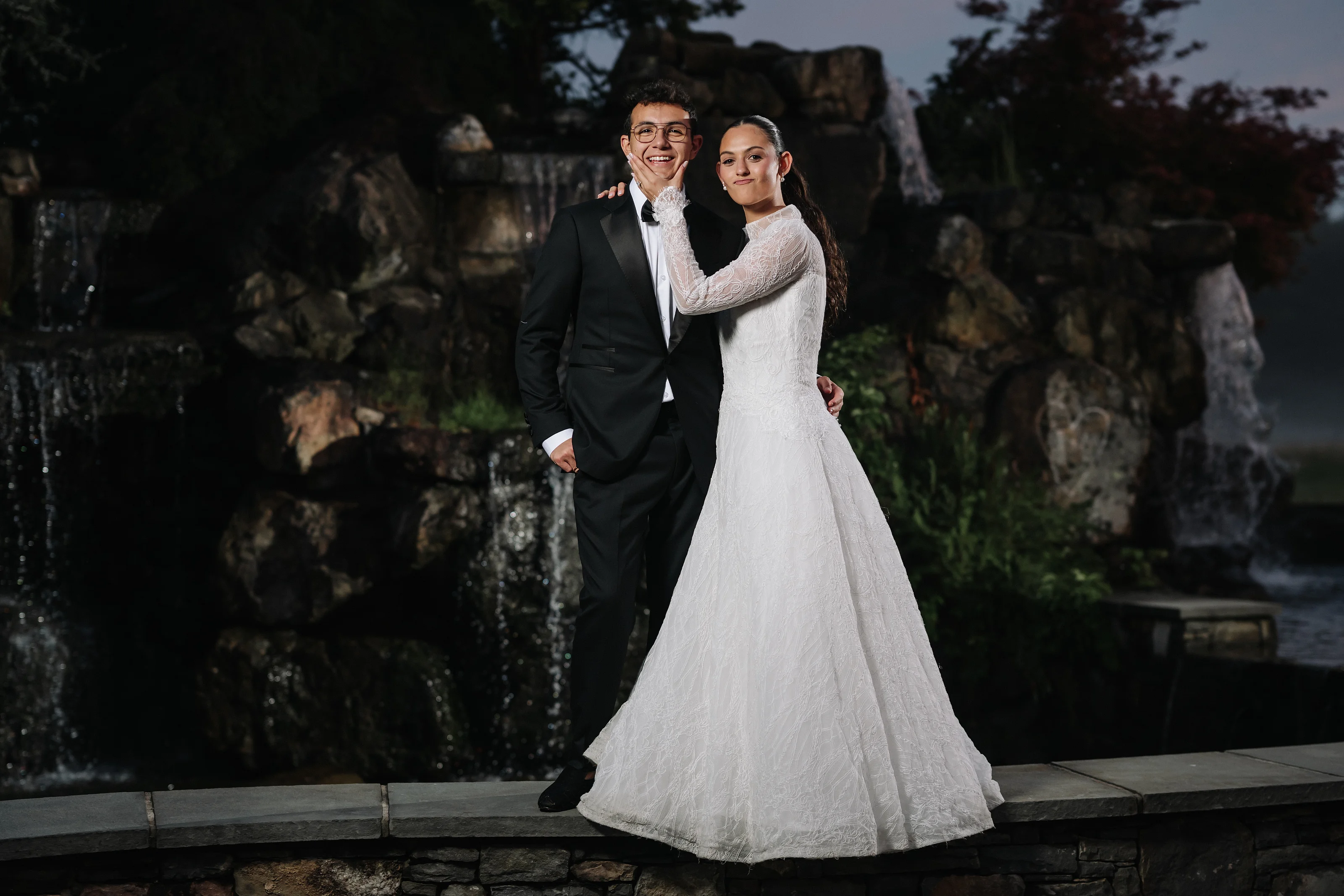 Bride and groom holding hands outdoors at dusk with a stone fountain backdrop, smiling as they walk forward.