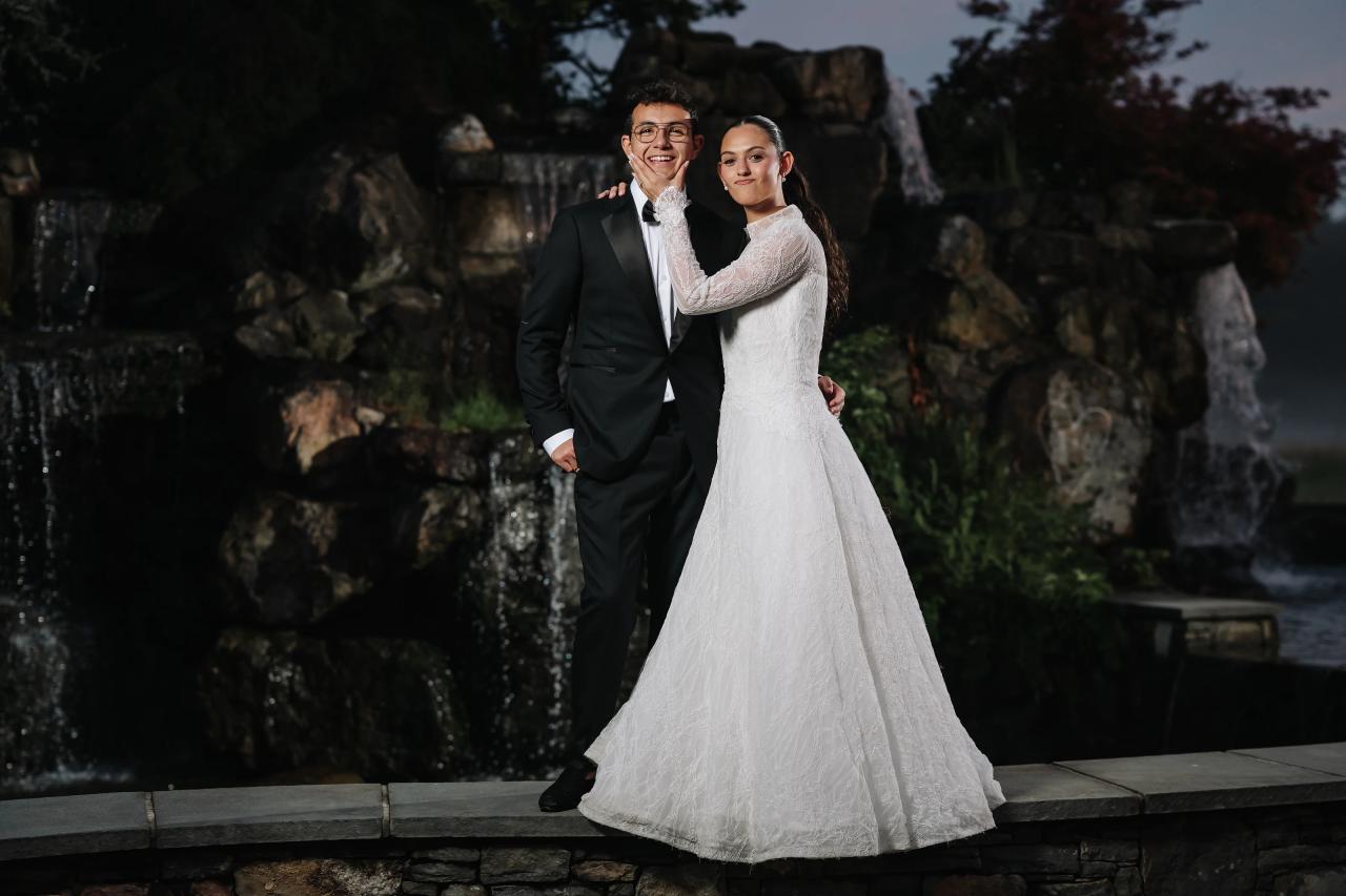 Bride and groom holding hands outdoors at dusk with a stone fountain backdrop, smiling as they walk forward.