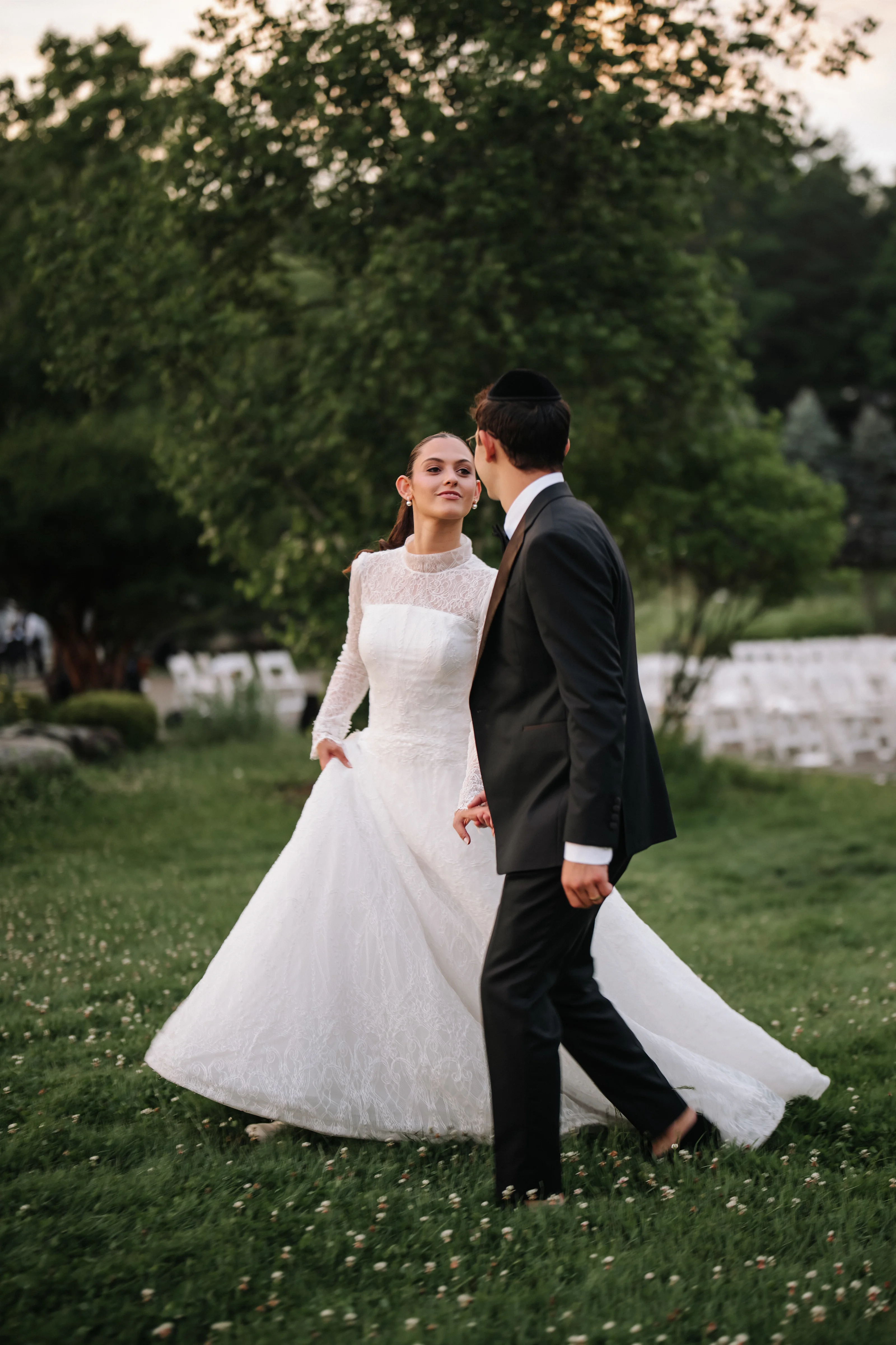 Bride and groom walking in a garden at sunset, bride looking over with veil and lace gown as groom faces her in a black suit.