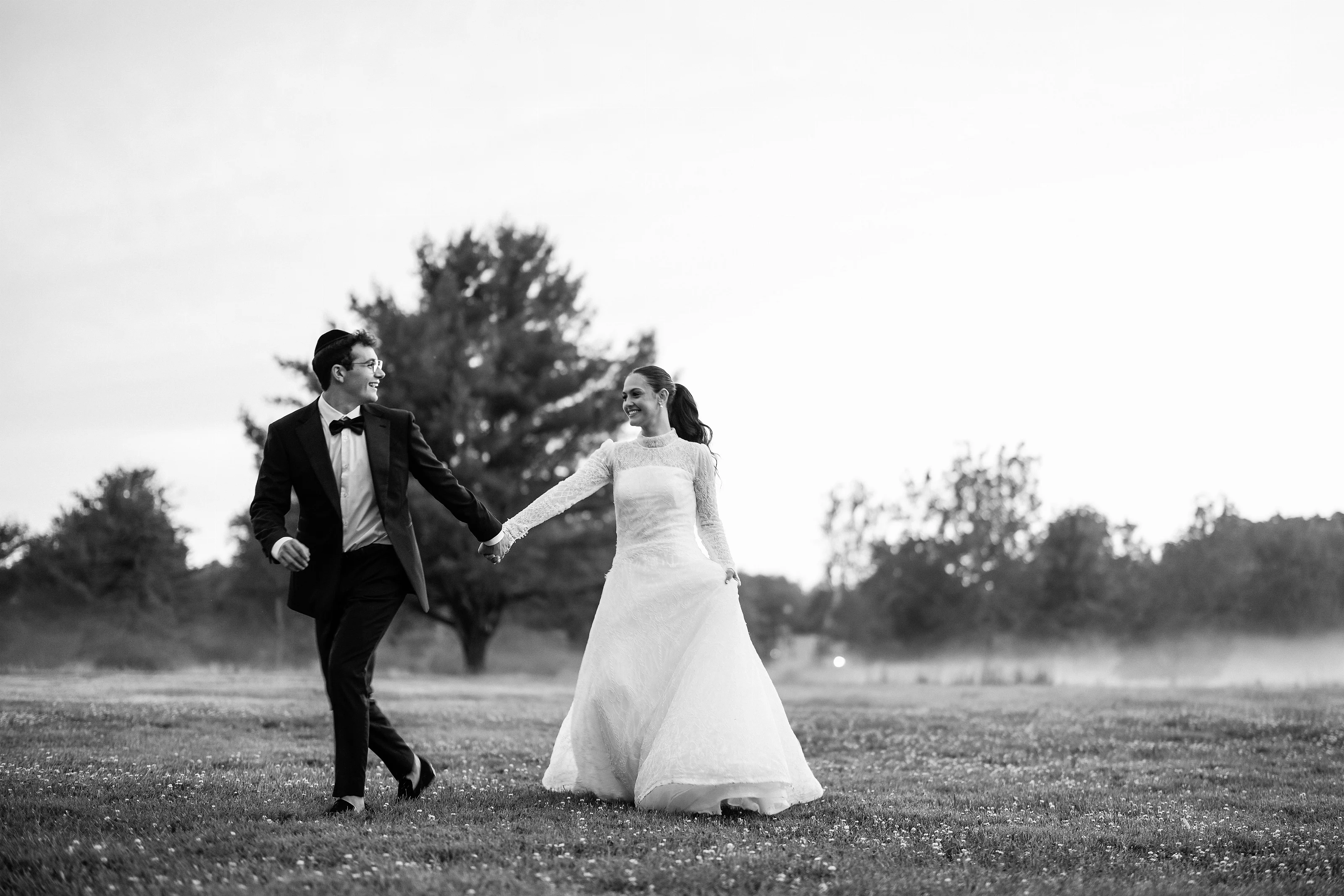 Black-and-white wedding photo of a bride and groom holding hands and walking across a field at sunset.