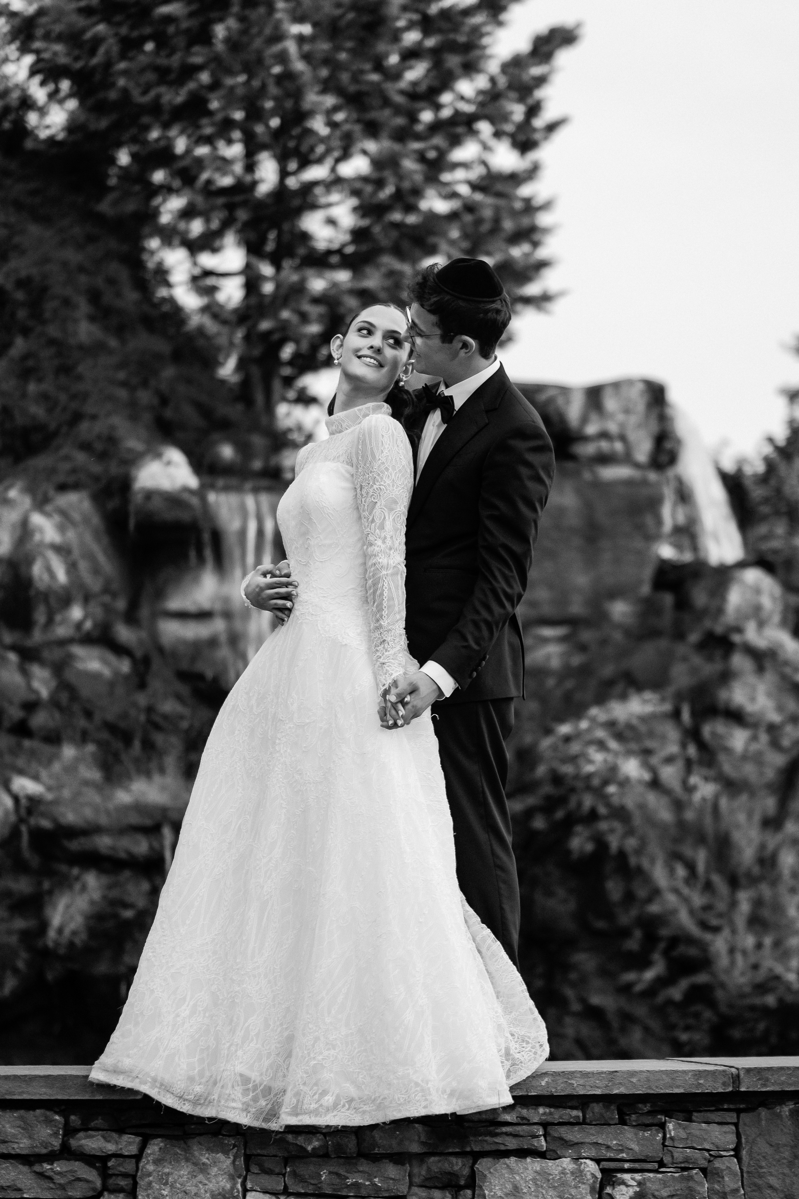 Black-and-white wedding portrait of a smiling bride and groom embracing outdoors, with a veil and lace dress in soft light.