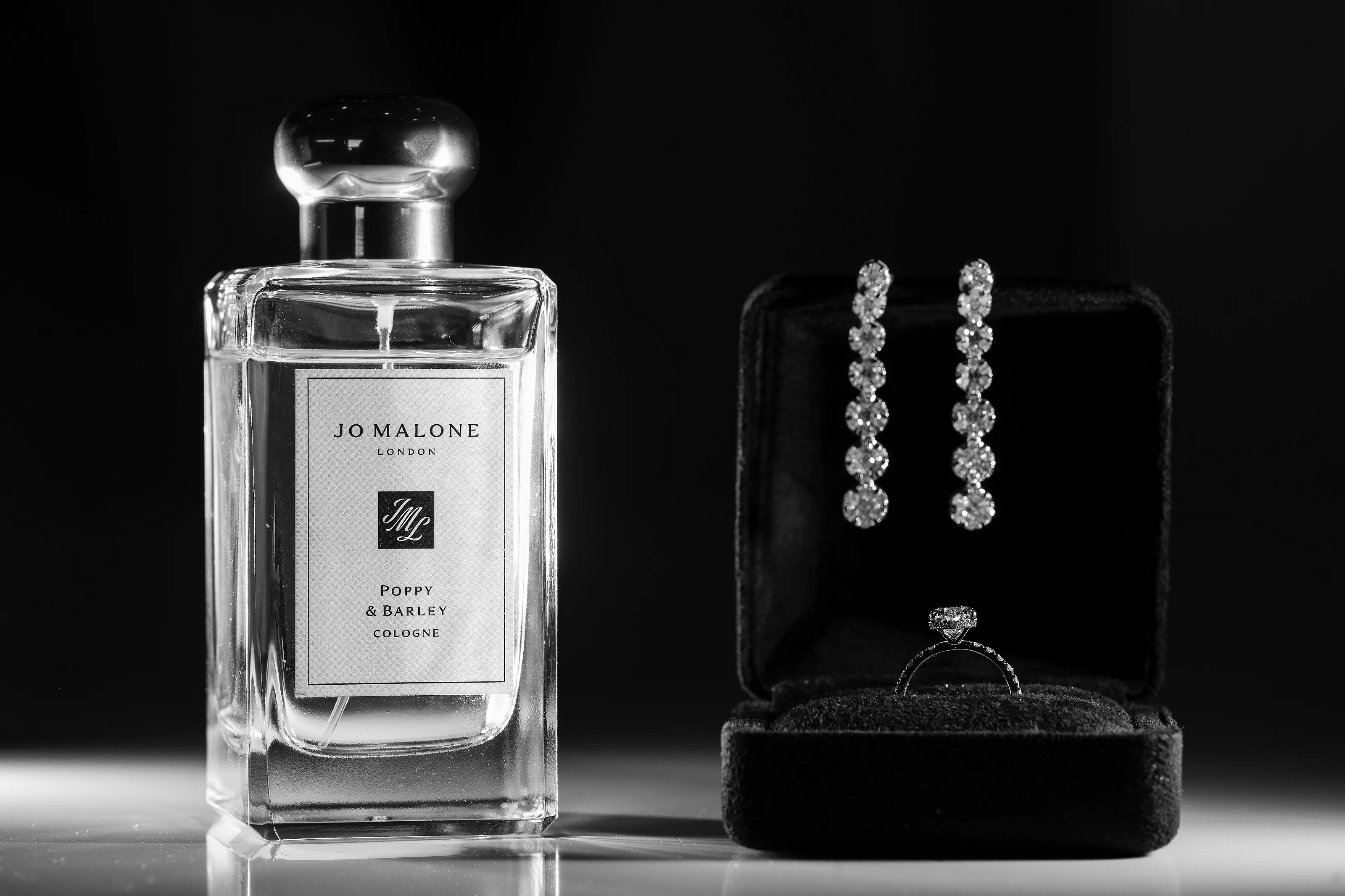 Black-and-white studio still life showing a perfume bottle beside diamond-like earrings on a dark ring box, all reflected on a glossy surface.