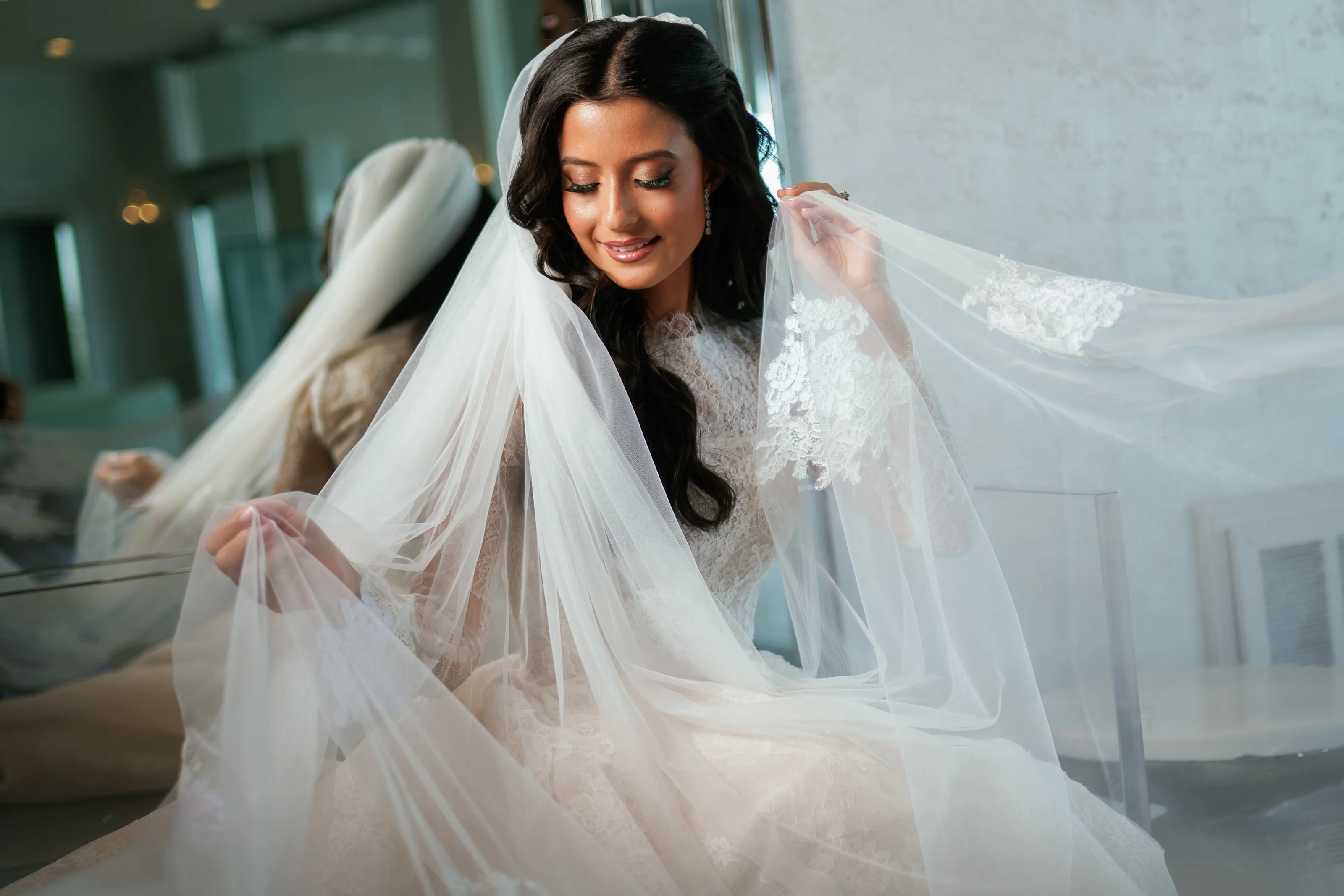 Bride in a lace gown with veil draped over her shoulders during badeken, gazing downward in soft window light.