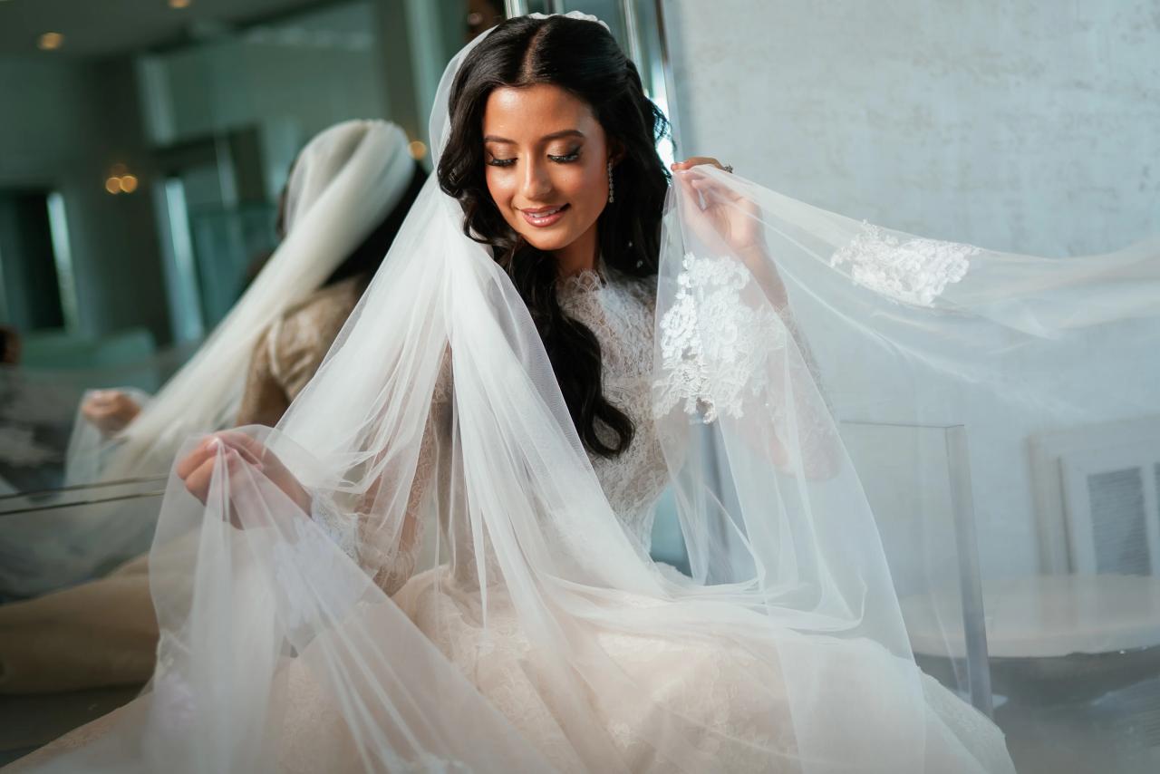 Bride in a lace gown with veil draped over her shoulders during badeken, gazing downward in soft window light. Wedding photography at Sands, Atlantic Beach, NY.