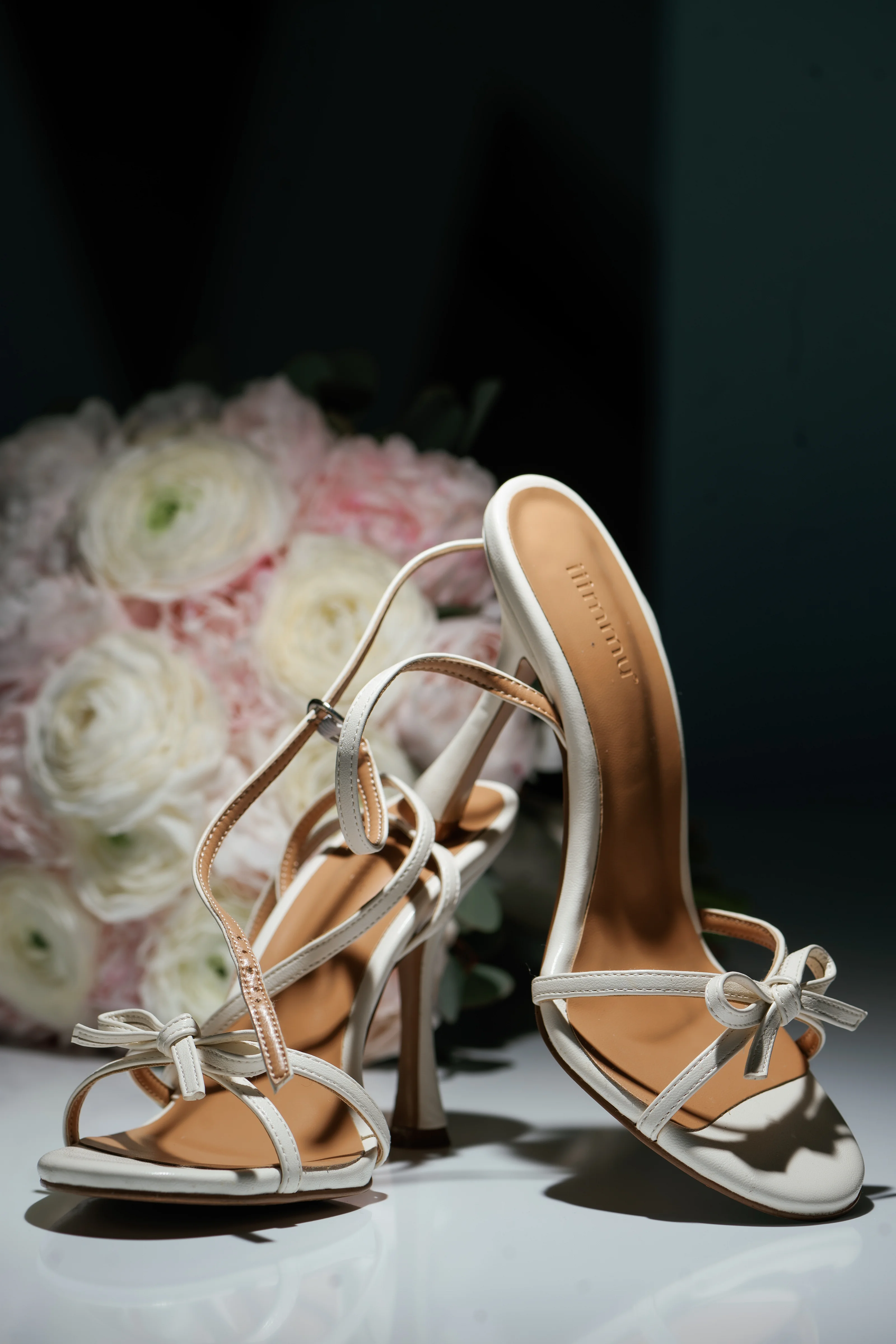 Close-up of white wedding heels with floral centerpiece softly blurred in the background.