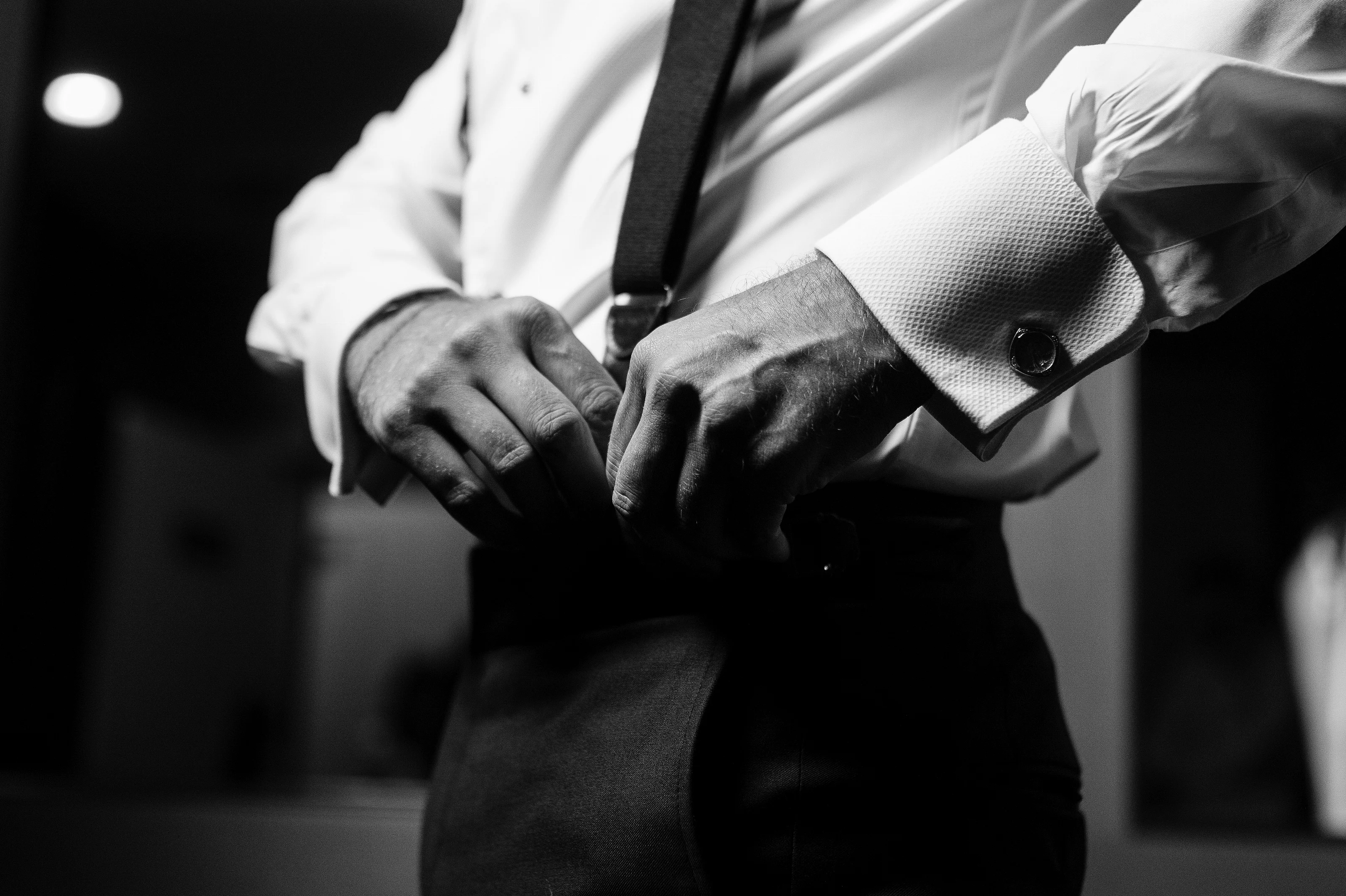 Black-and-white close-up of a groom’s hands adjusting and fastening tefillin, with focused texture on the prayer garment.