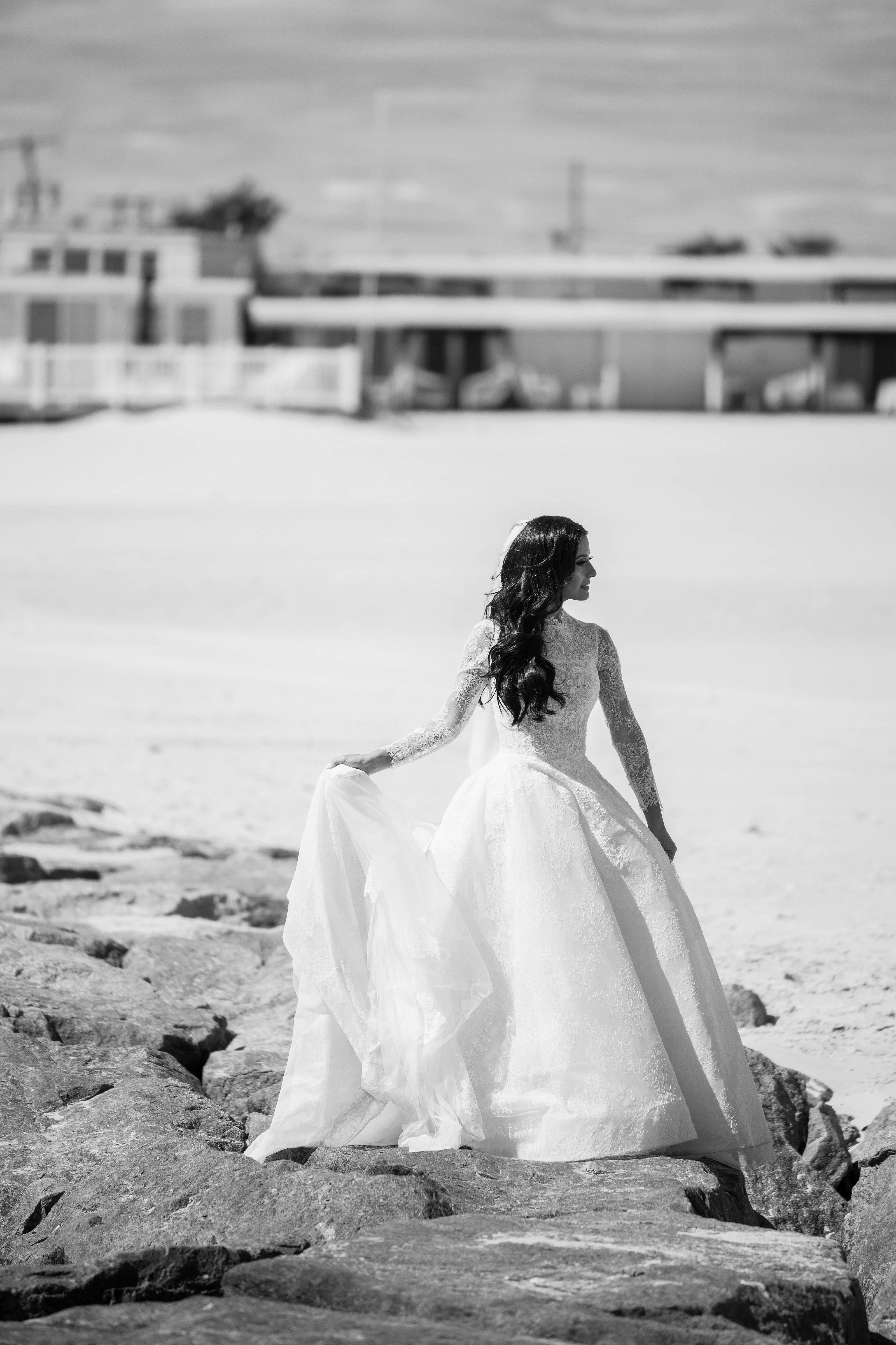 Black-and-white photo of a bride walking across rocky ground outdoors, her gown and veil catching the light as she turns in profile.