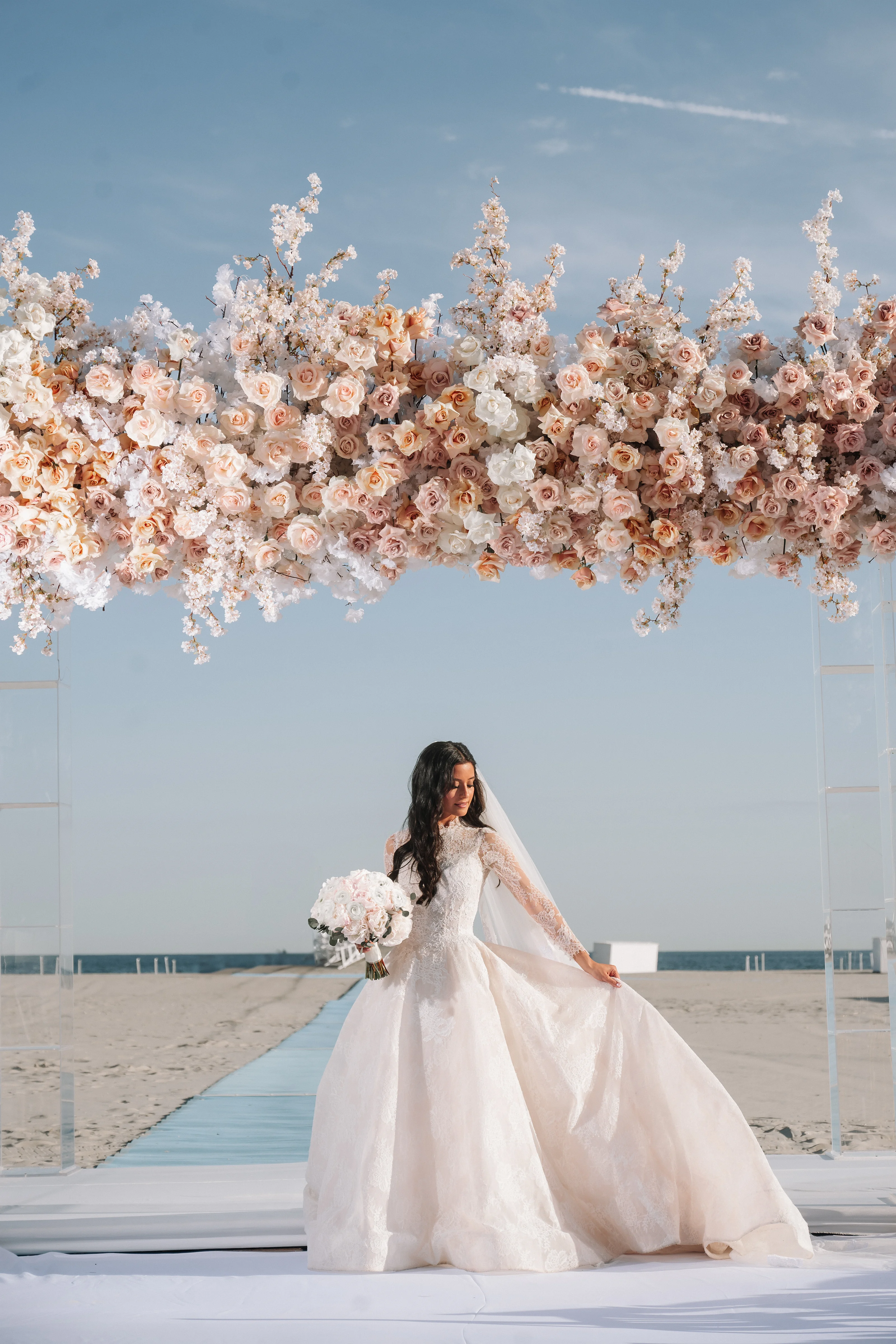 Bride in a white gown and veil holding a bouquet on a beach walkway beneath an overhead installation of white and blush flowers against a blue sky.