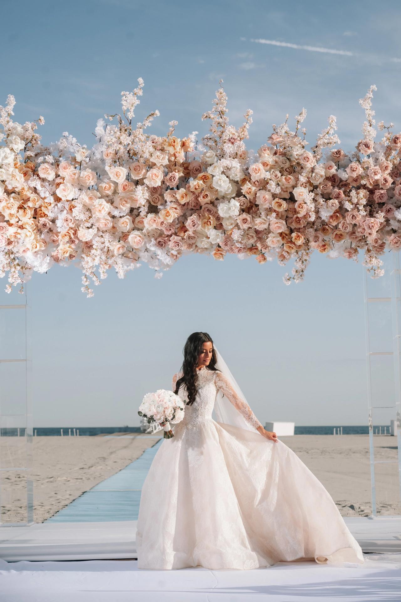 Bride in a white gown and veil holding a bouquet on a beach walkway beneath an overhead installation of white and blush flowers against a blue sky. Wedding photography at Sands, Atlantic Beach, NY.
