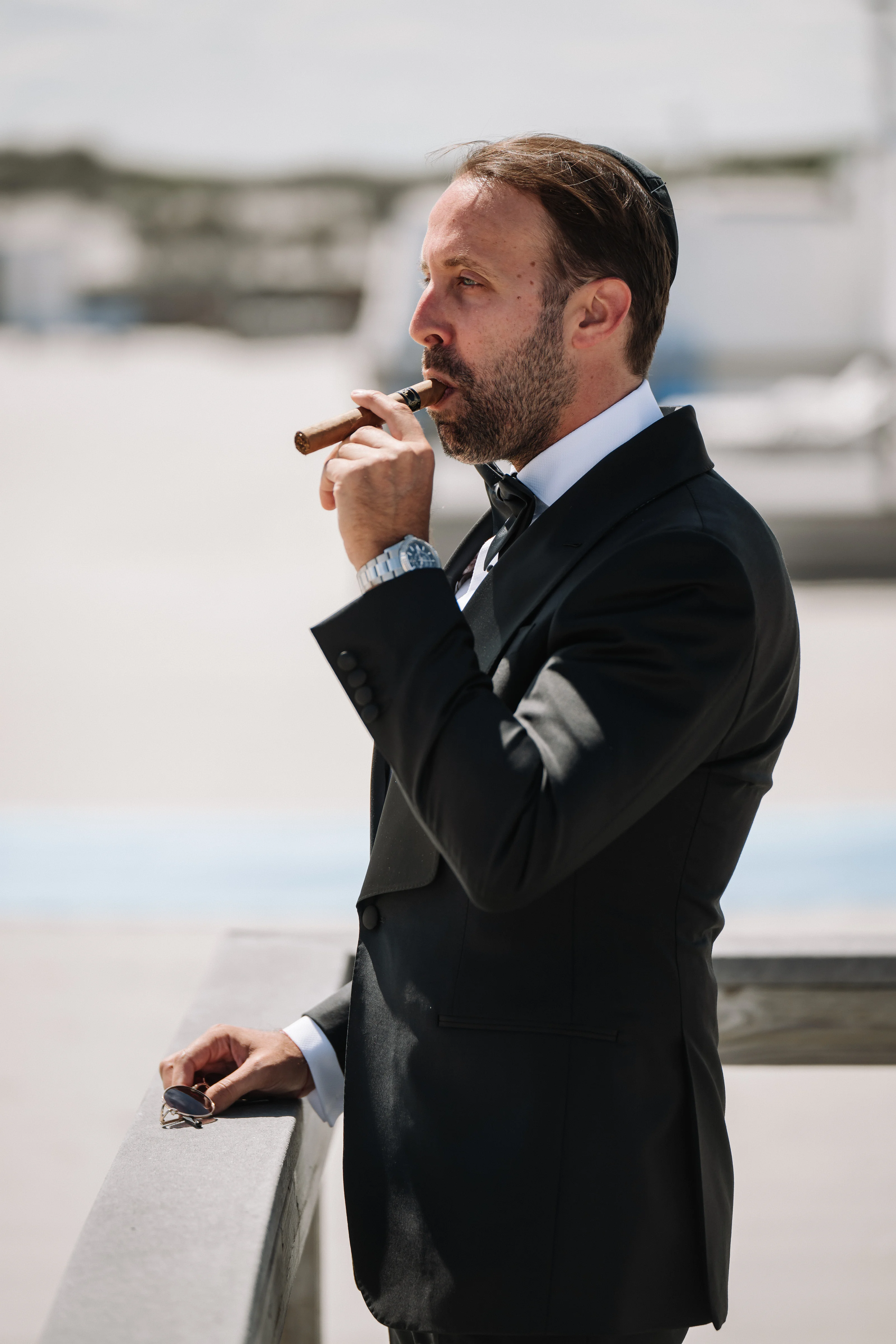 Groom in a dark suit outdoors in side profile holding a cigarillo near his mouth, caught in a focused moment with shallow depth of field.