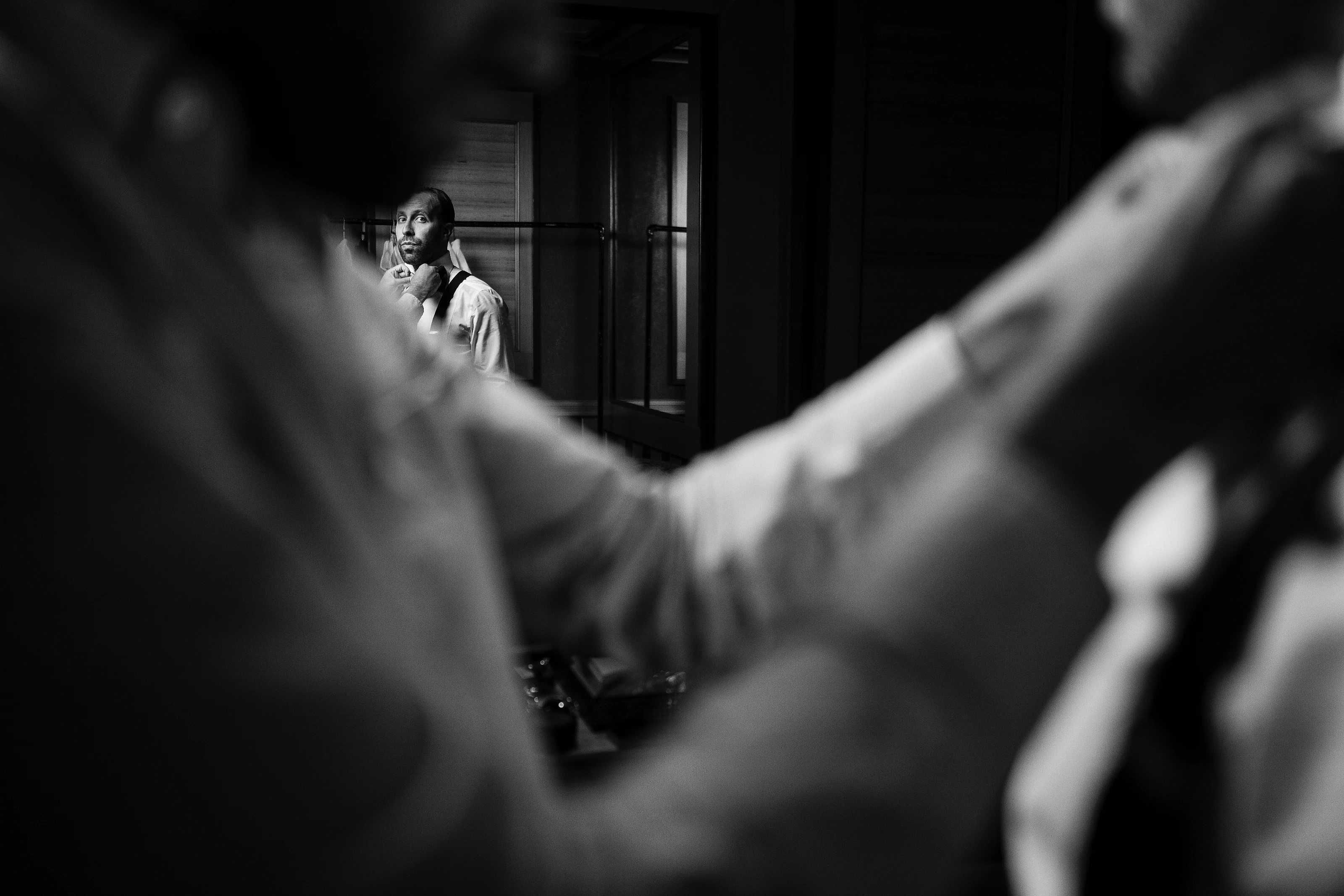 Black-and-white wedding moment framed through blurred foreground arms, with the groom holding a drink visible in the background hallway.
