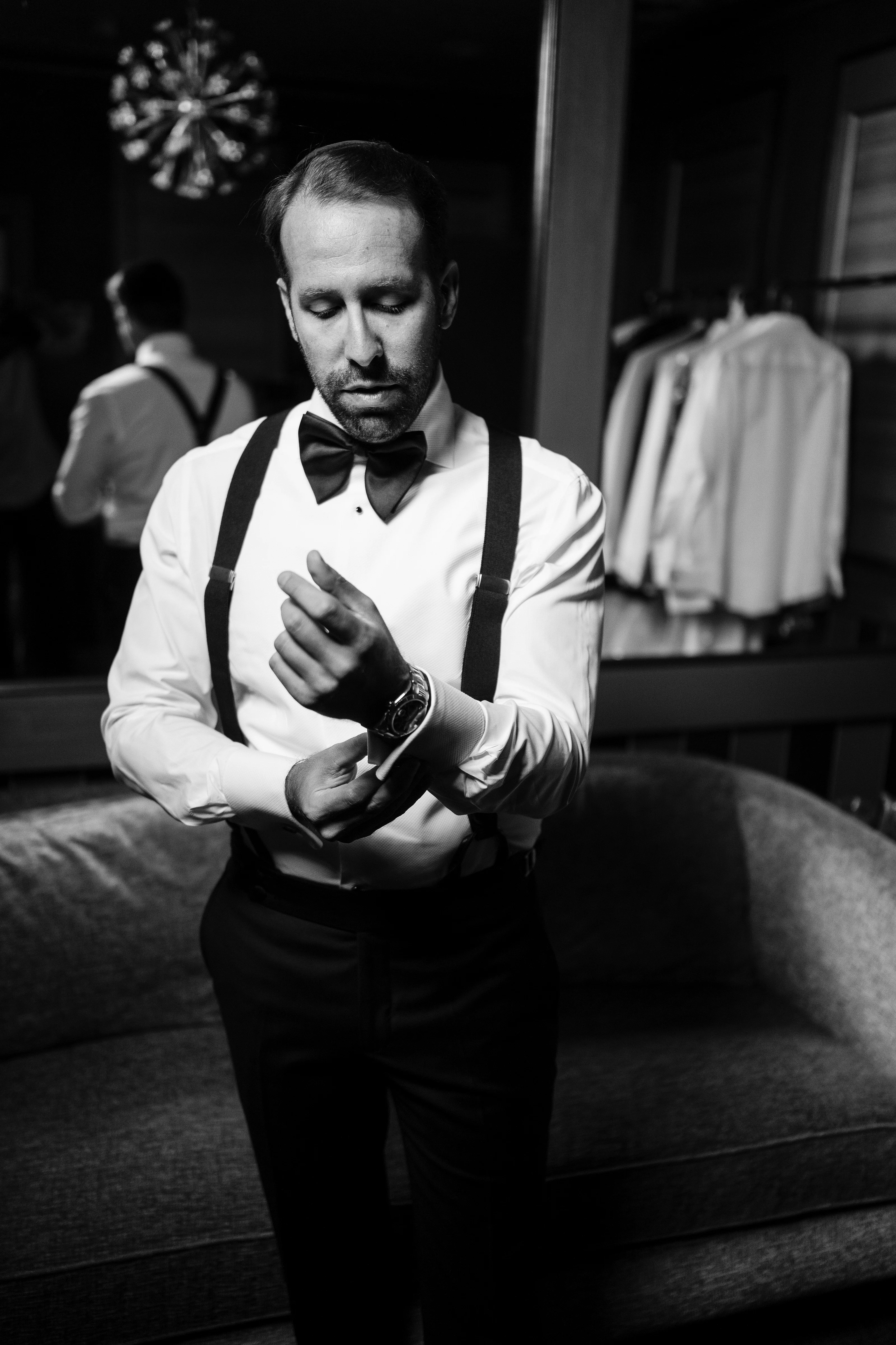 Black-and-white photo of a groom in a tuxedo and bow tie with suspenders, hands raised in a quiet moment during the reception, guests softly blurred in the background.