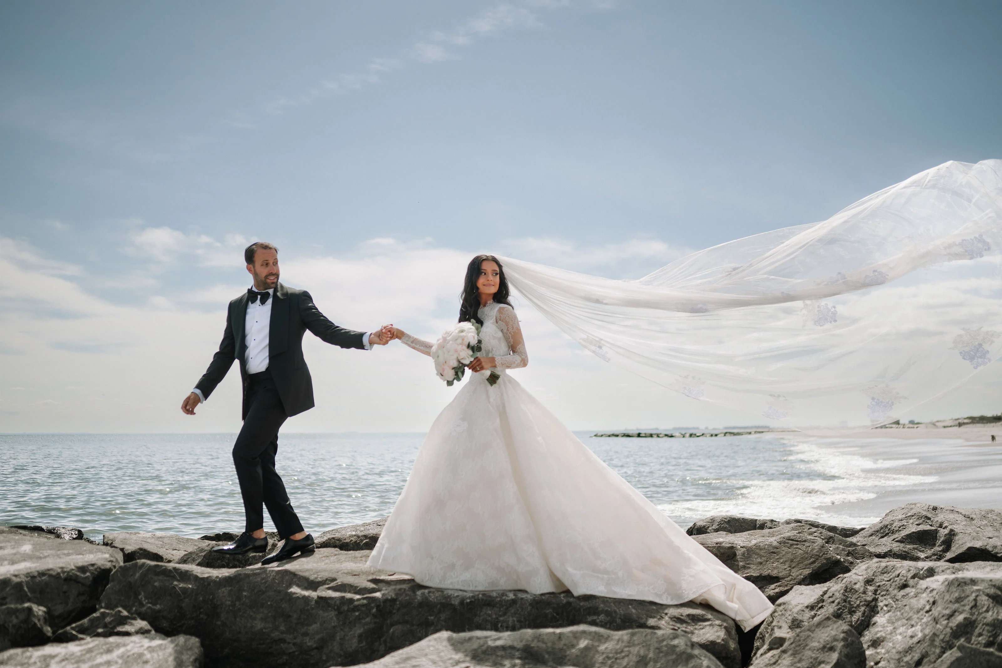 Bride and groom holding hands on rocky coastline as a long veil billows in the wind with ocean and mountains in the background.