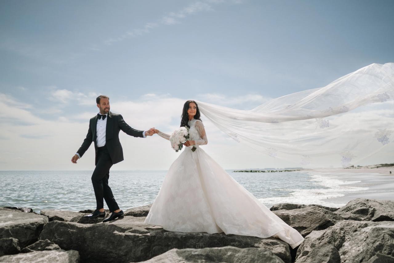 Bride and groom holding hands on rocky coastline as a long veil billows in the wind with ocean and mountains in the background. Wedding photography at Sands, Atlantic Beach, NY.