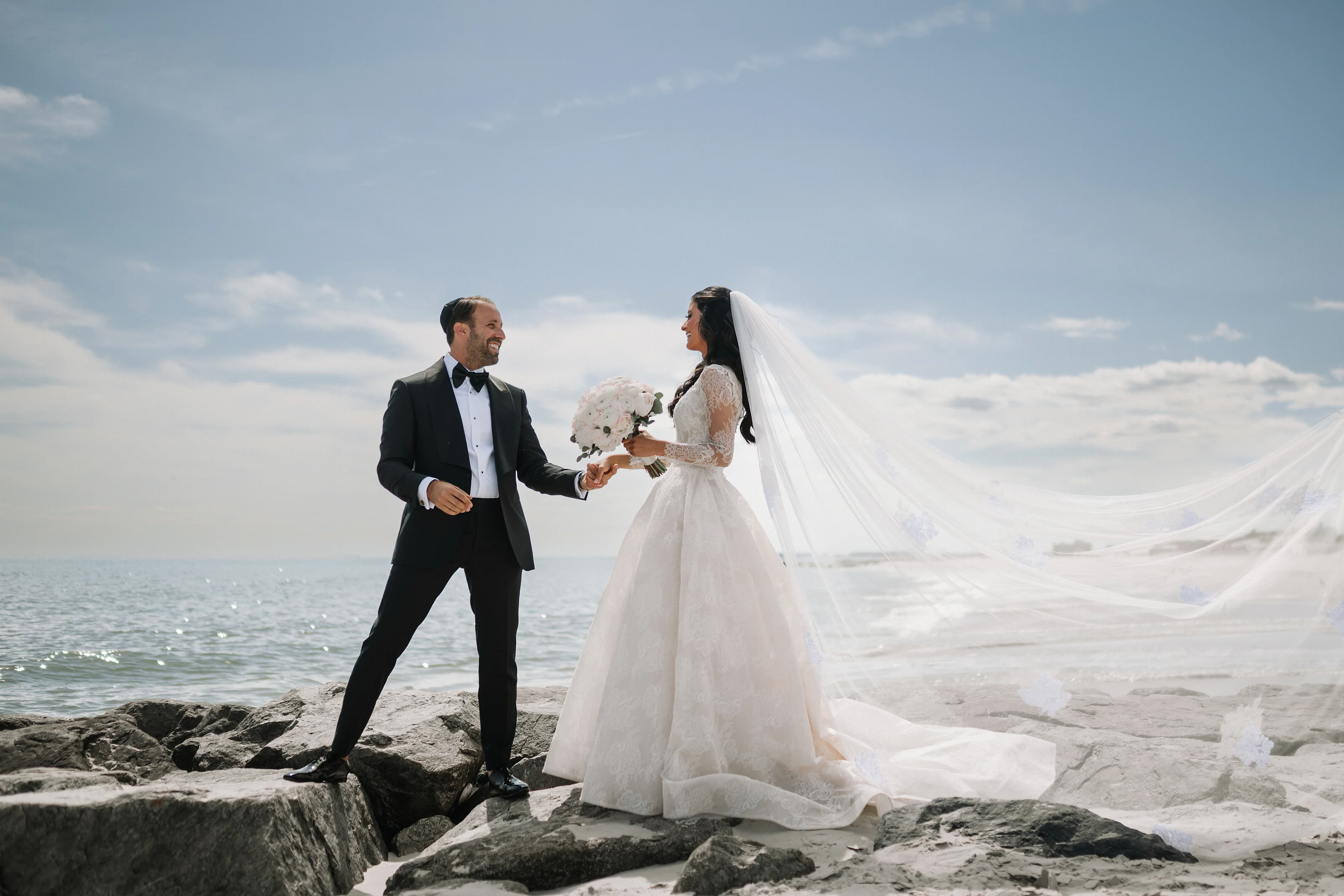 Bride in a white gown and veil walking beside groom on a rocky shoreline, holding a bouquet as they smile at each other under a cloudy sky.