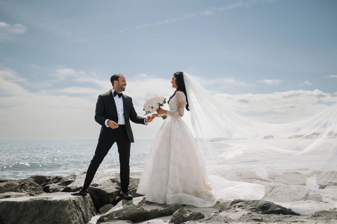 Bride in a white gown and veil walking beside groom on a rocky shoreline, holding a bouquet as they smile at each other under a cloudy sky. Wedding photography at Sands, Atlantic Beach, NY.