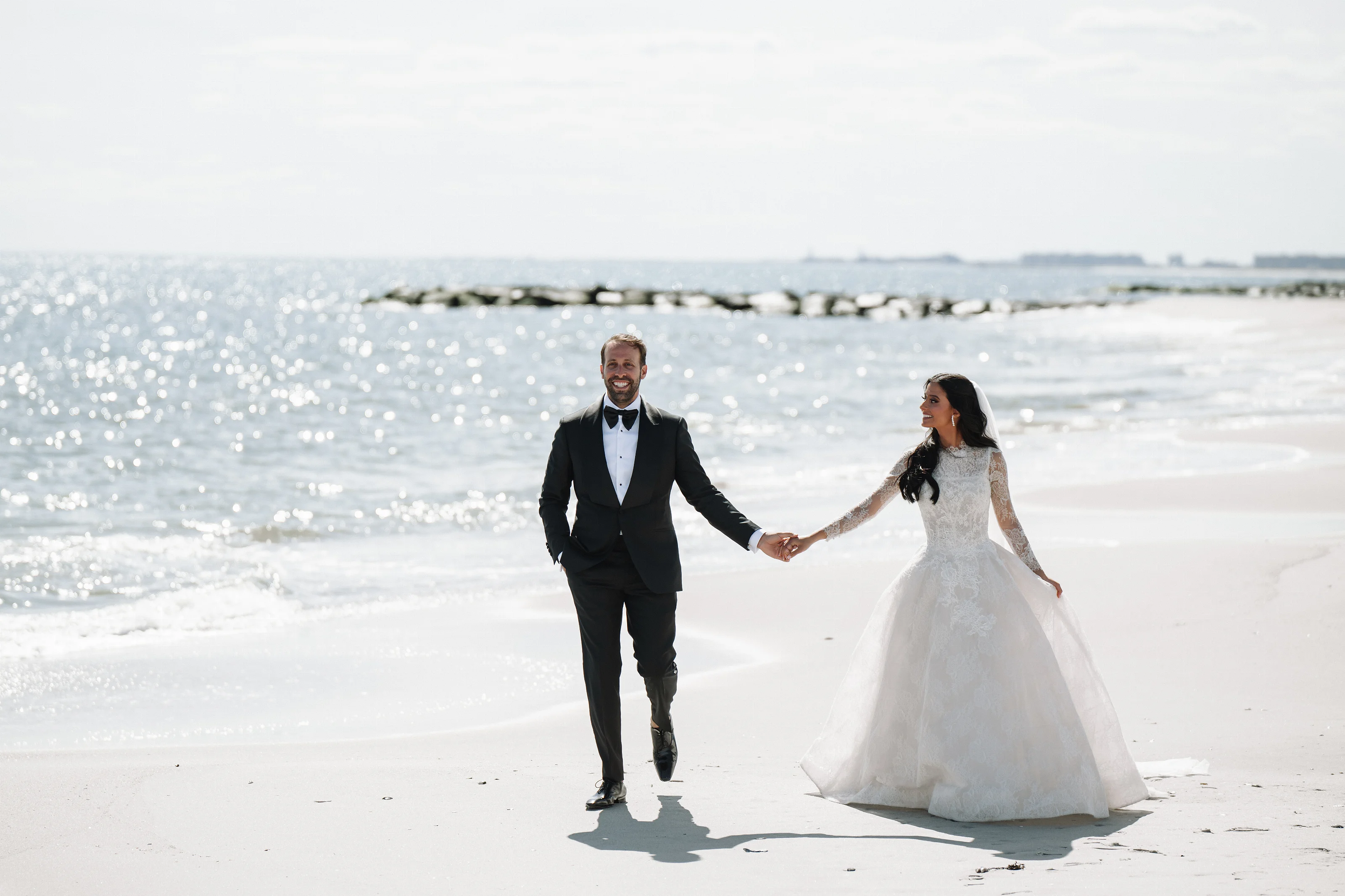 Bride and groom holding hands while walking along a sunny beach with the ocean and shoreline in the background.