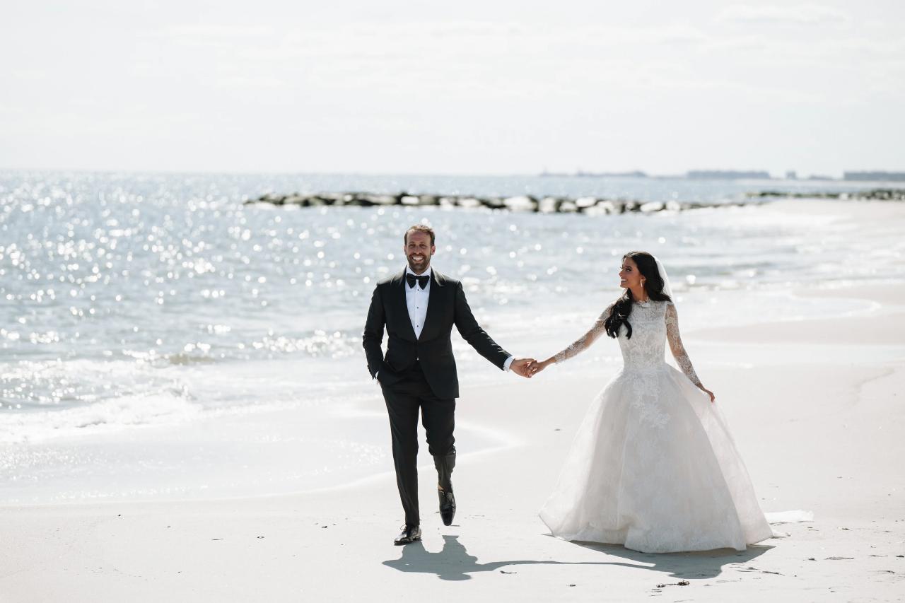 Bride and groom walking hand in hand along a bright beach at The Sands in Atlantic Beach, New York.