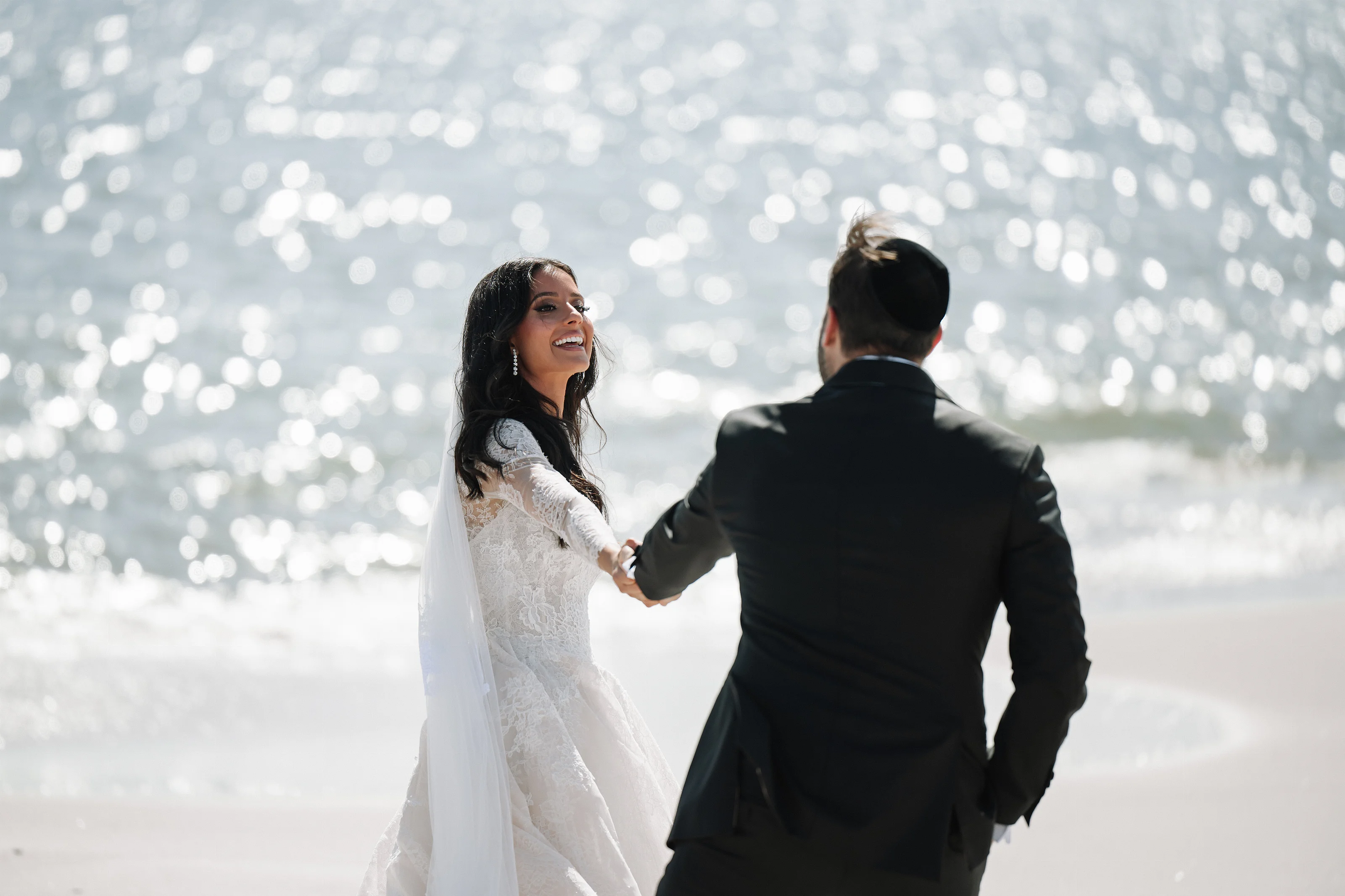 Bride in a white veil and groom in dark formalwear walking hand in hand, smiling with a bright bokeh light backdrop.