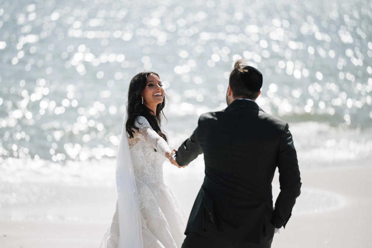 Bride in a white veil and groom in dark formalwear walking hand in hand, smiling with a bright bokeh light backdrop. Wedding photography at Sands, Atlantic Beach, NY.