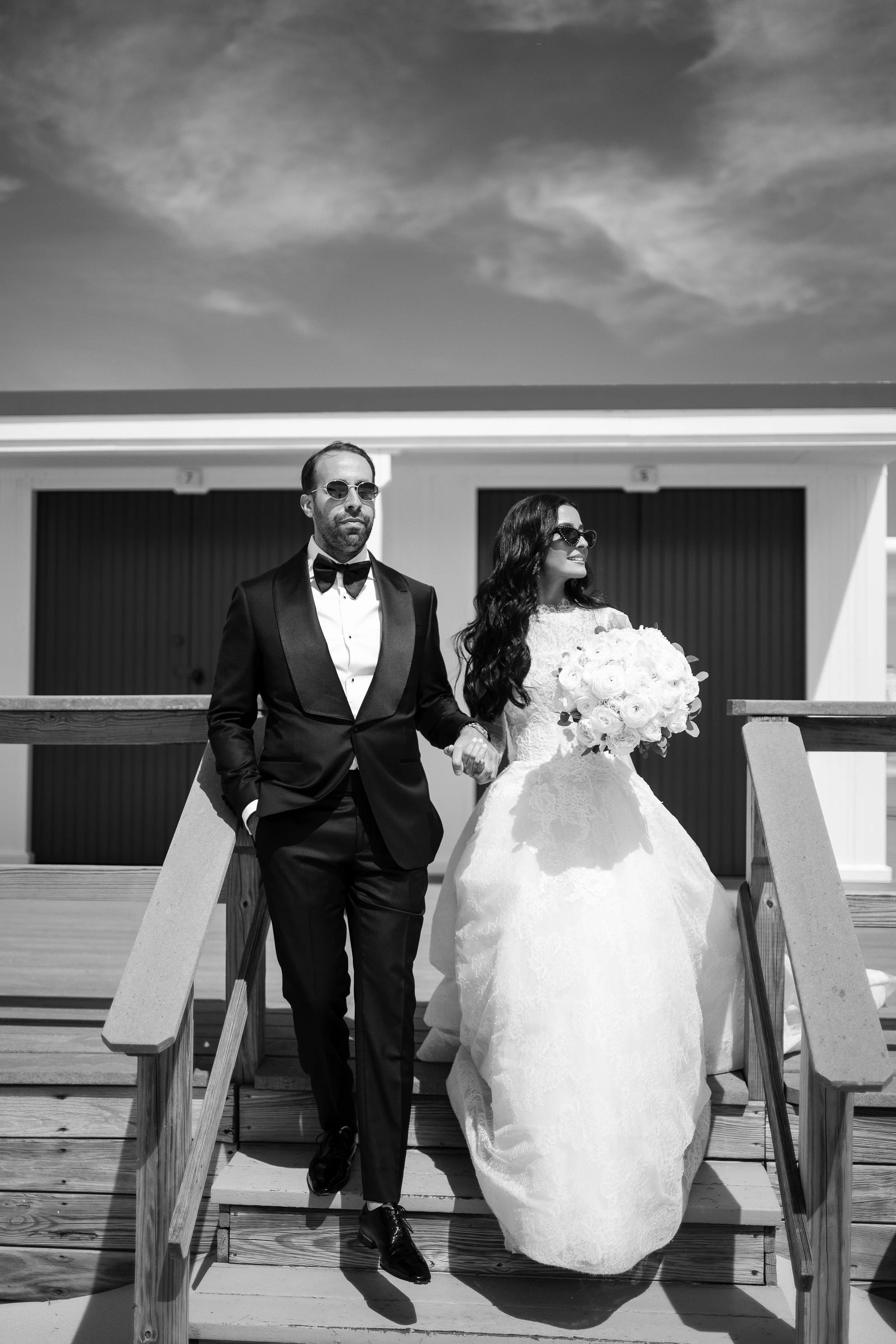 Black-and-white photo of a bride and groom walking hand-in-hand down outdoor steps, holding hands and looking off to the side.