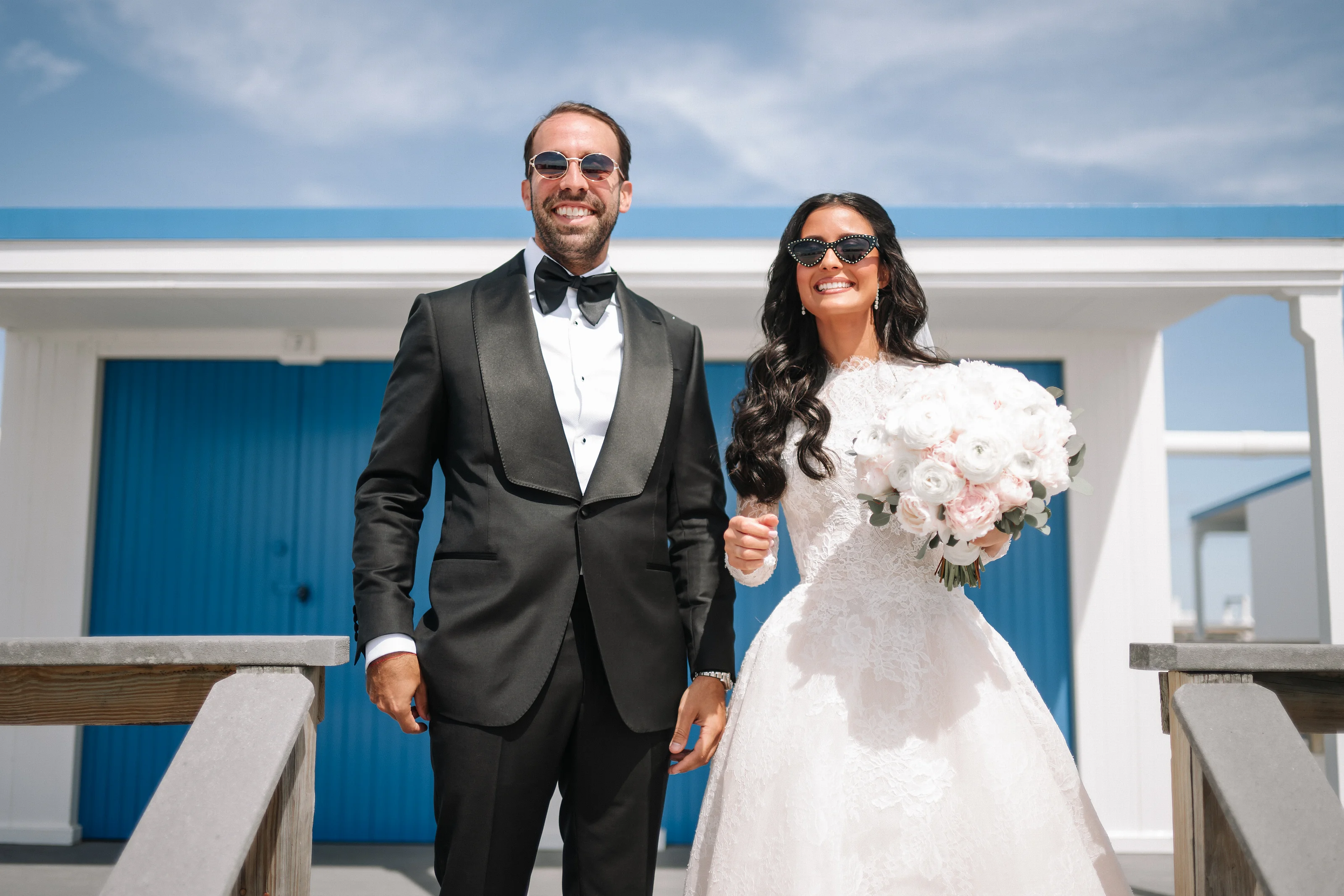 Wedding couple smiling and walking together hand-in-hand outdoors, bride holding a white bouquet with the ocean visible in the background.