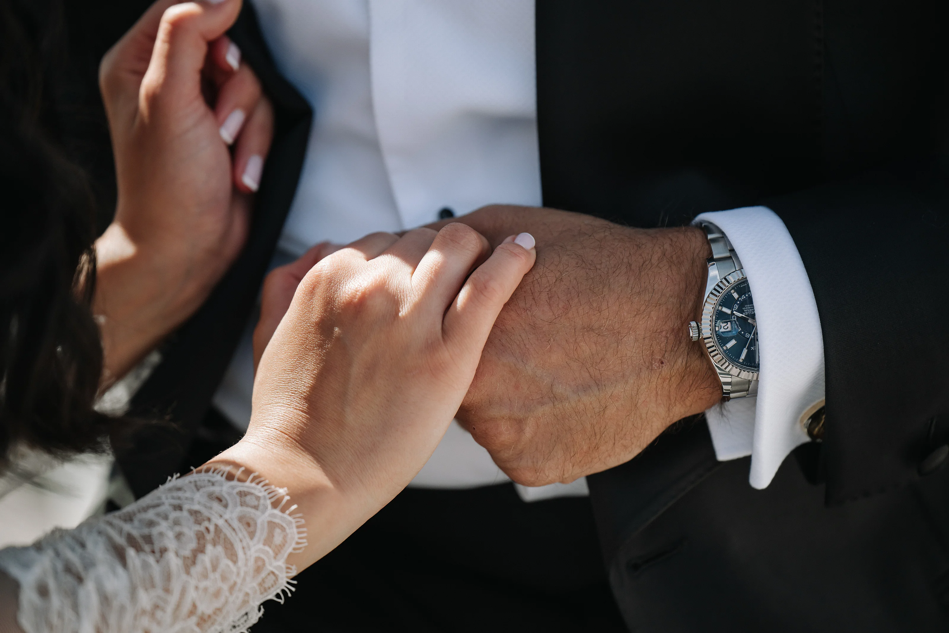 Close-up of a bride’s hands touching the groom’s wrist during a wedding ritual, with a watch visible and soft warm light on skin.