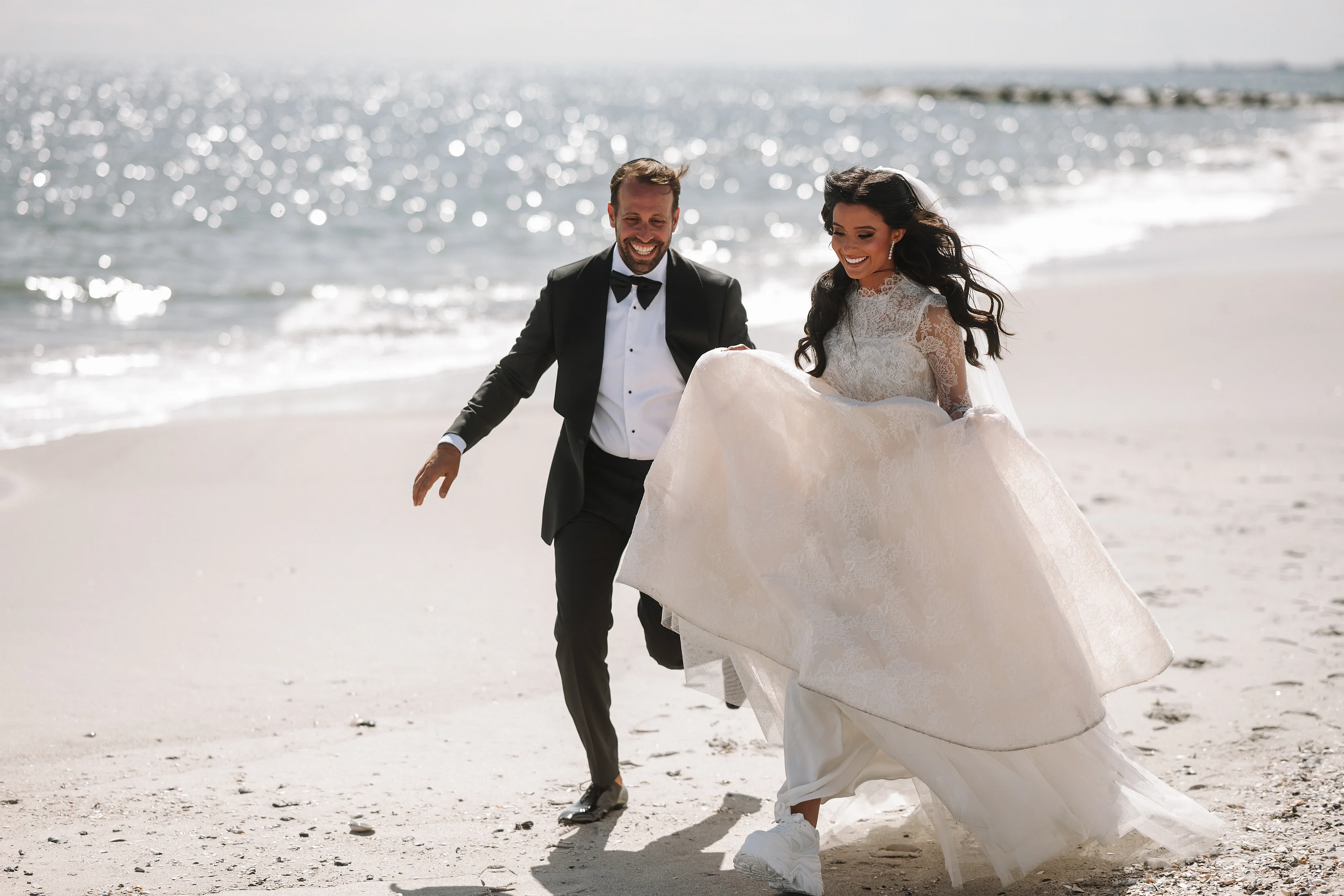 Bride and groom walking together on a sunny beach, bride’s dress billowing, ocean sparkle in the background.