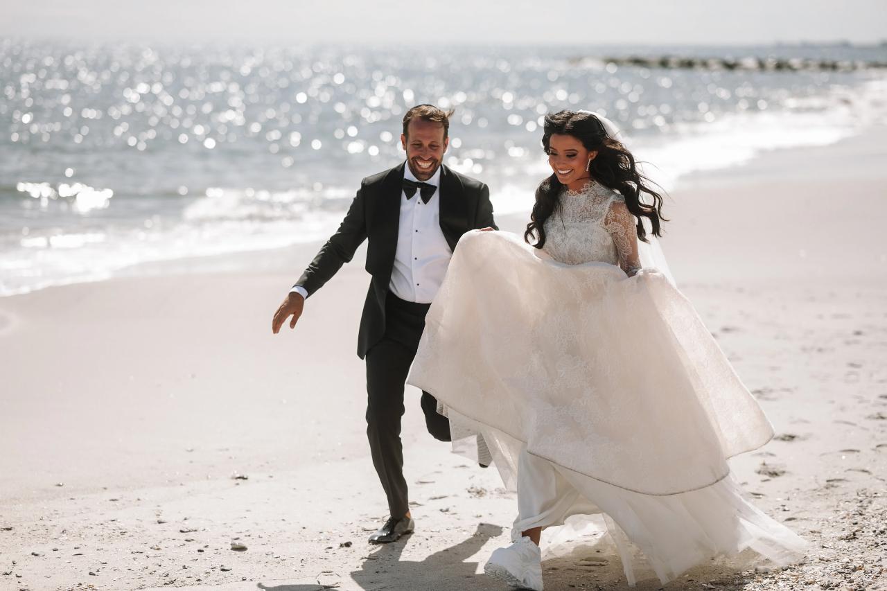 Bride and groom walking together on a sunny beach, bride’s dress billowing, ocean sparkle in the background.