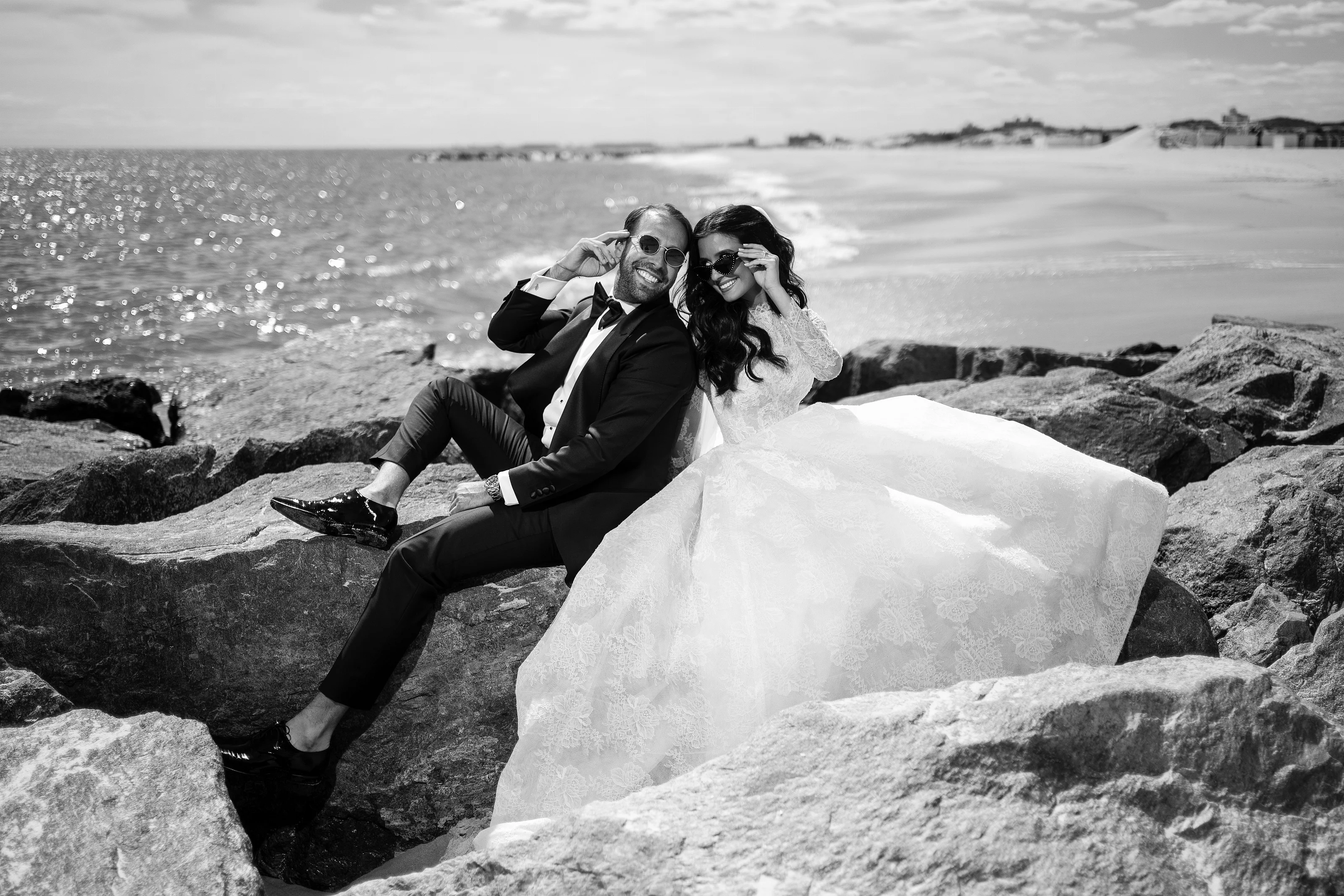 Black-and-white seaside wedding portrait of a laughing bride and groom seated on rocks with ocean waves sparkling behind them.
