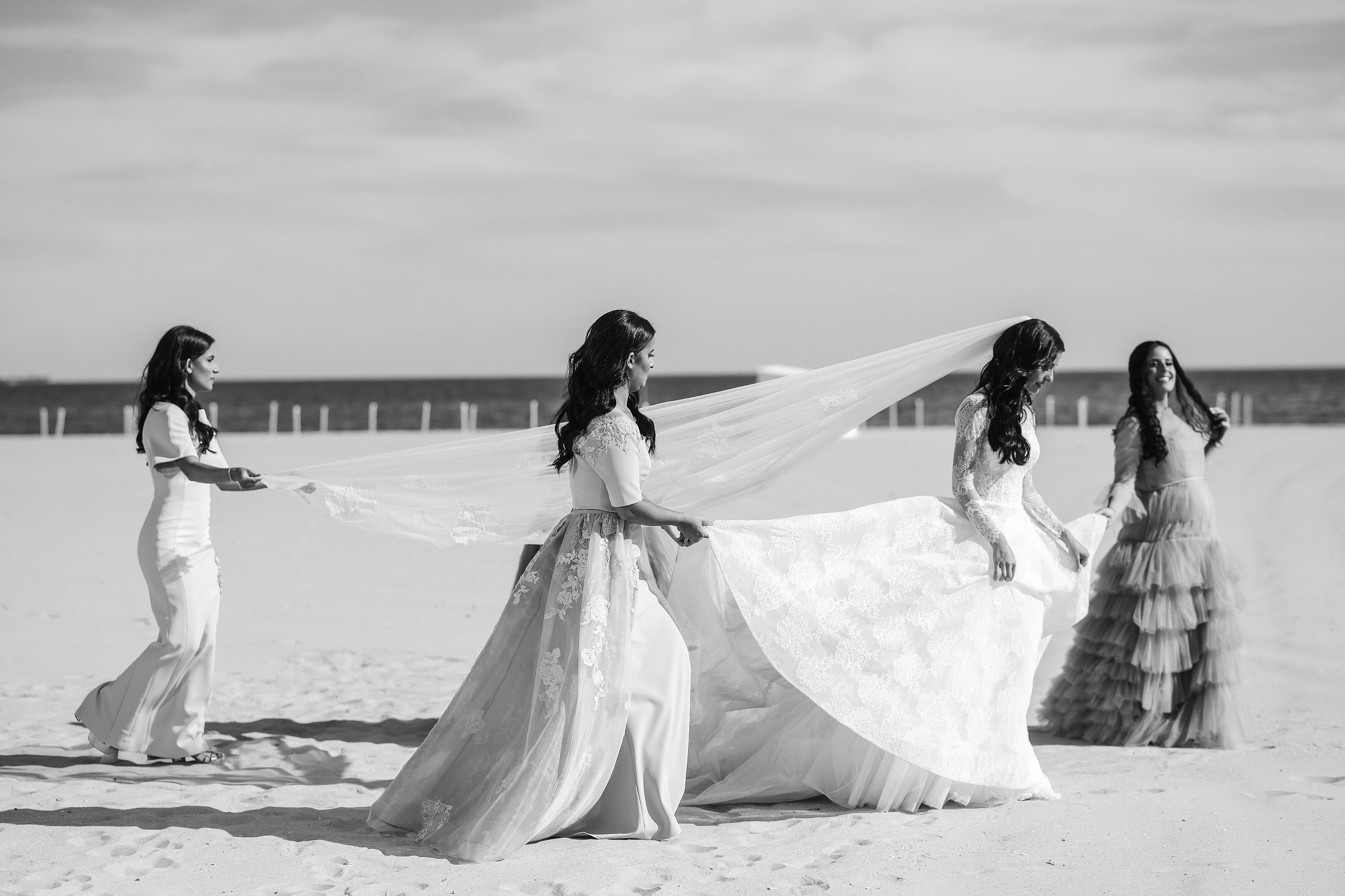 Black-and-white beach photo of a bride in a flowing gown with her veil billowing as she moves forward, surrounded by friends in the background.