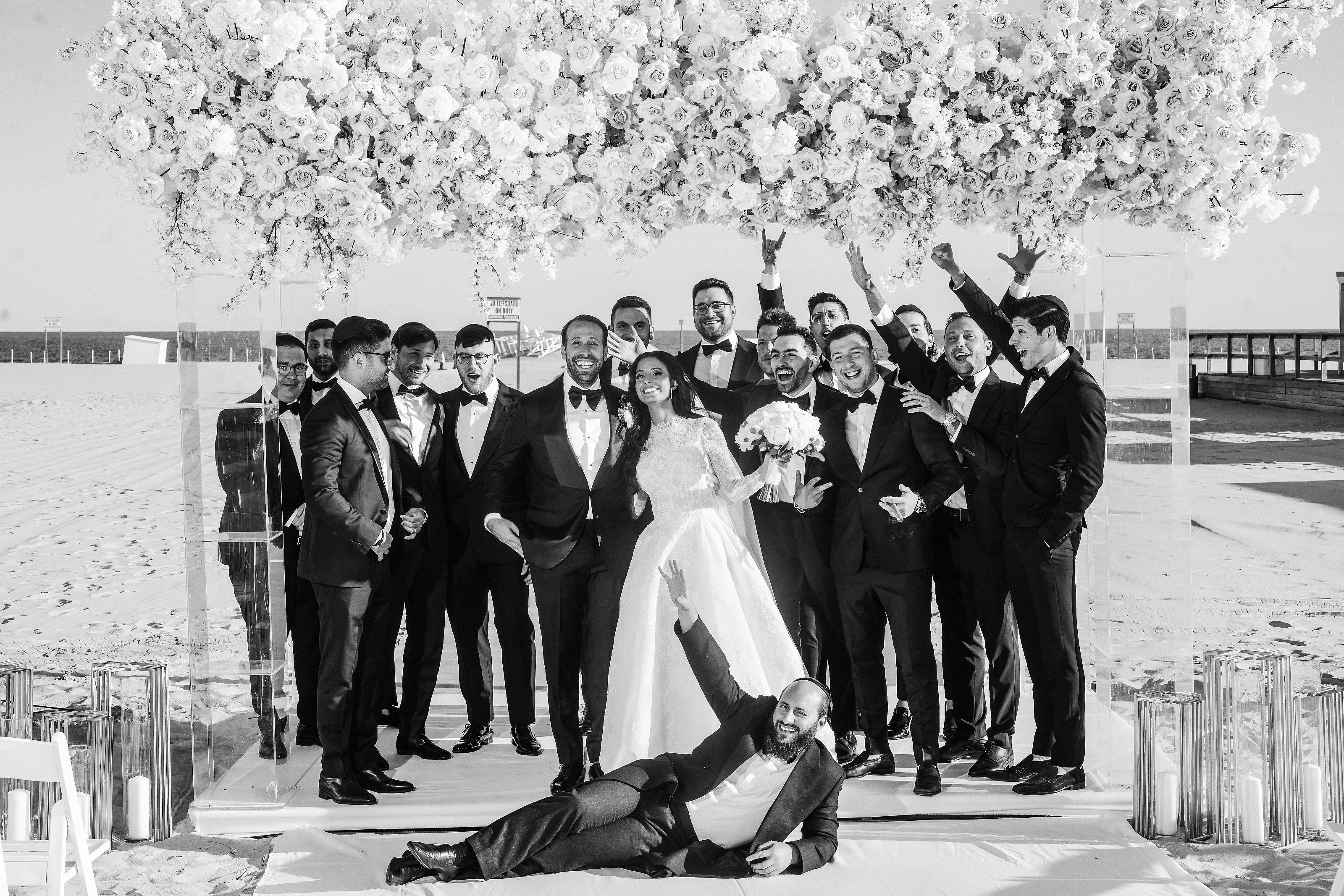 Black-and-white group celebration at a wedding ceremony, with the bride centered on stage, the groom reclining in the foreground, and guests in tuxedos smiling and raising their arms under a floral canopy.