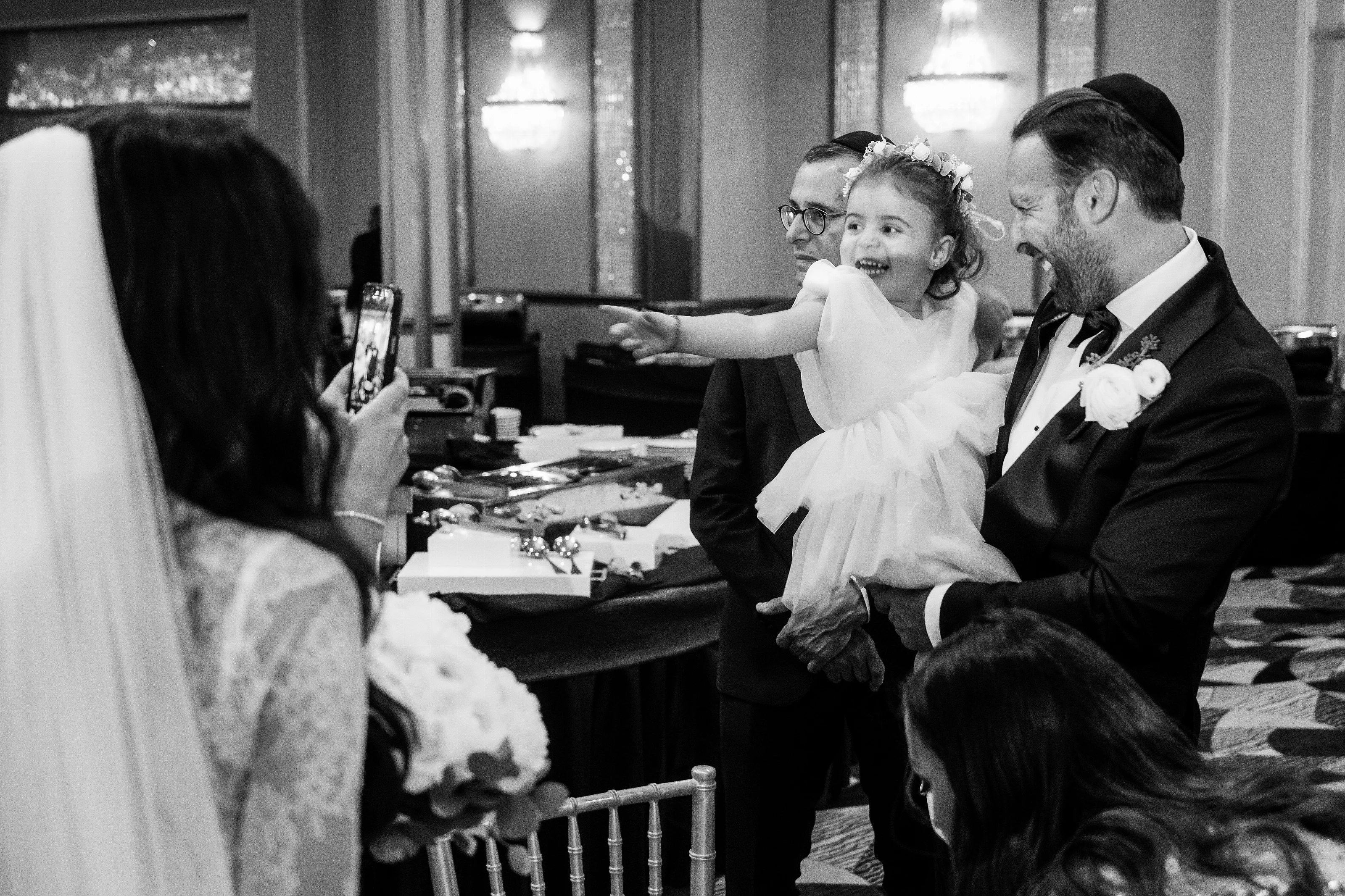 Black-and-white candid wedding moment of a bride and groom smiling as they hold their laughing child during a reception.