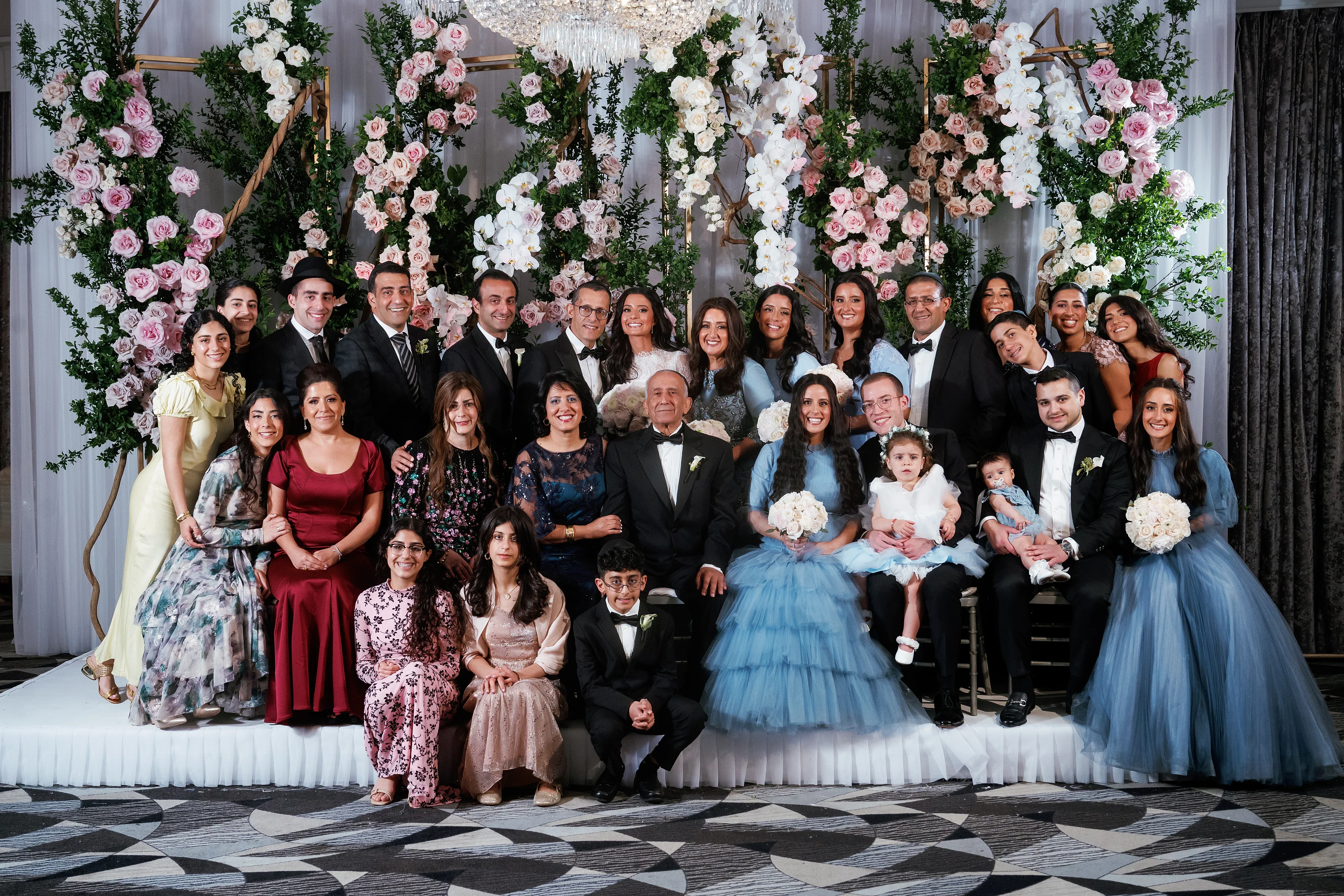 Large group family portrait from a wedding or mitzvah, posed in front of an elaborate floral backdrop with greenery and pastel flowers.