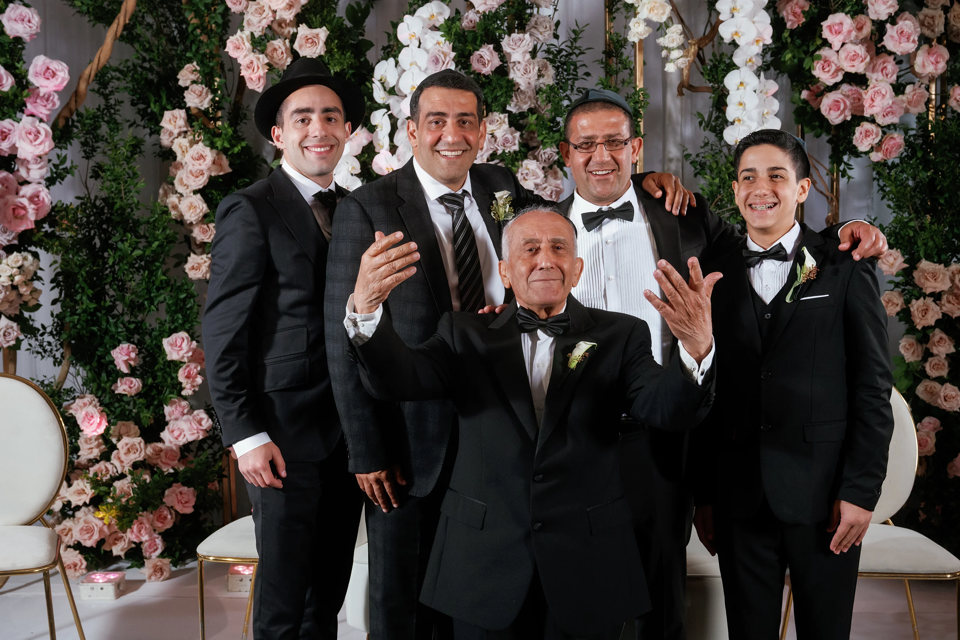 Smiling family members in black formal attire pose together in front of a pink and white floral backdrop, with one seated man centered and surrounded by others.