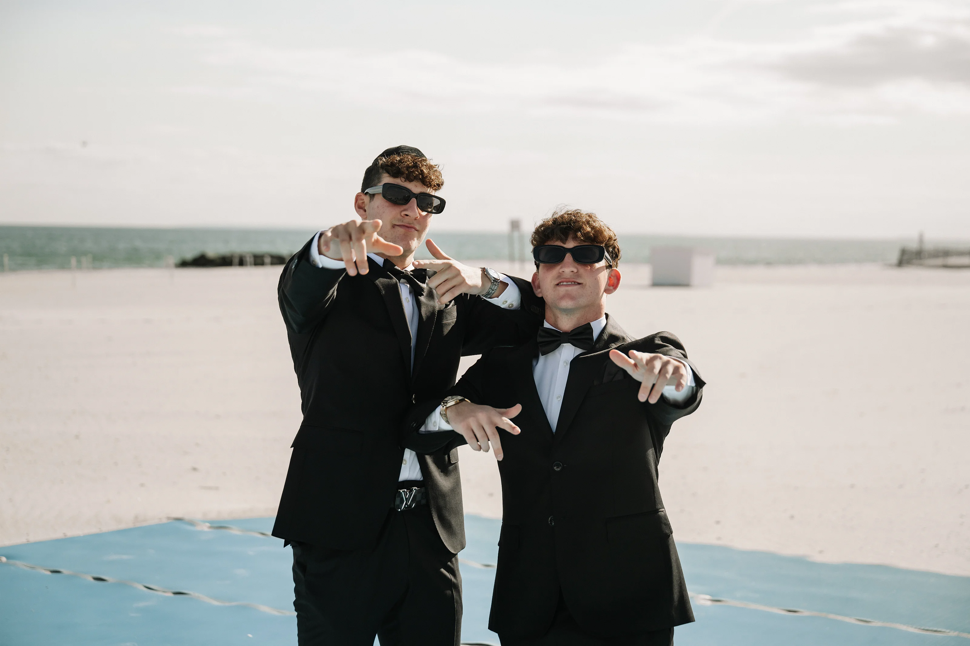 Two men in black-tie outfits at a beach wearing sunglasses and making peace signs toward the camera, with the ocean and distant shoreline behind them.
