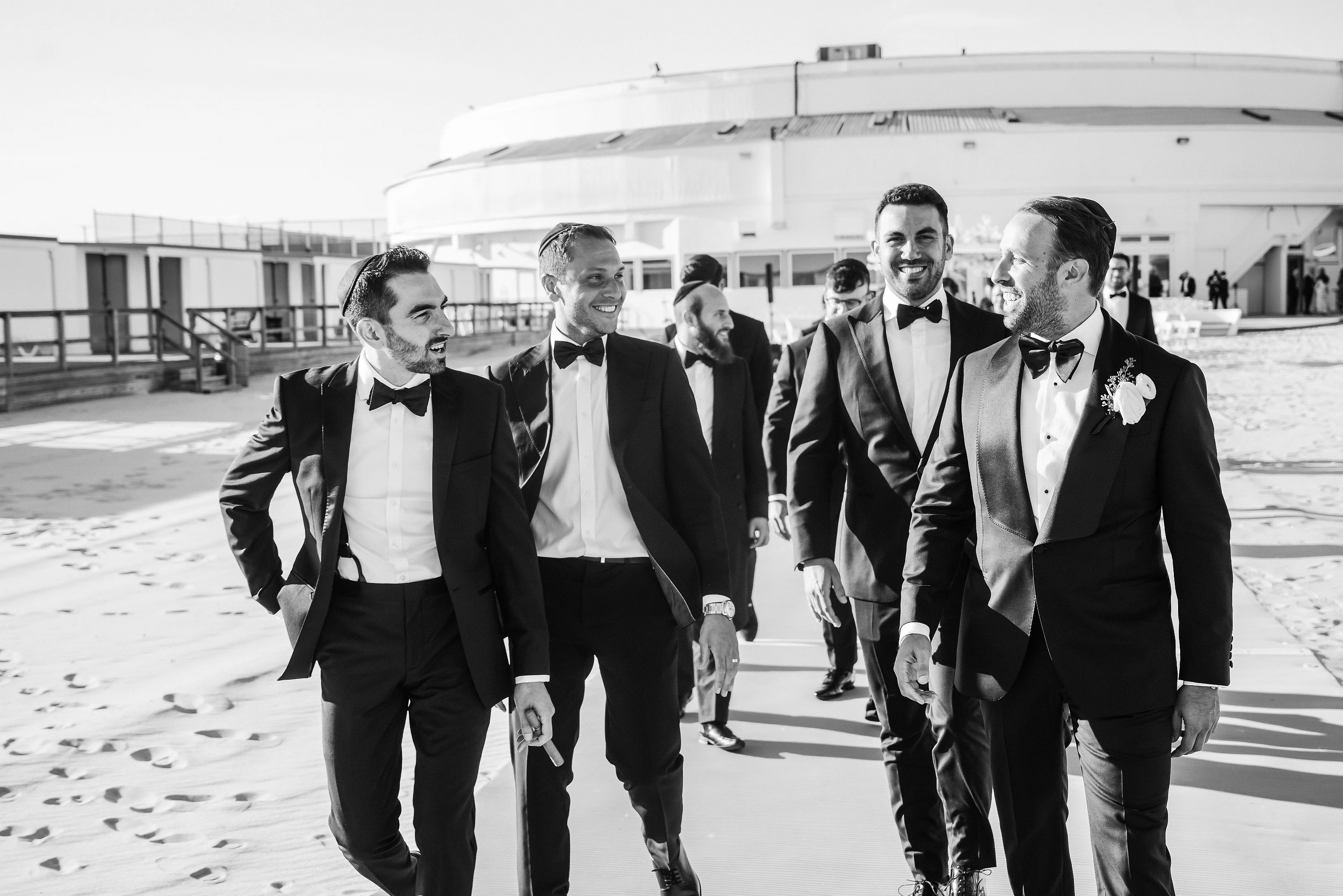 Black-and-white photo of the groom in black-tie walking with close family and smiling outdoors with a large building in the background.