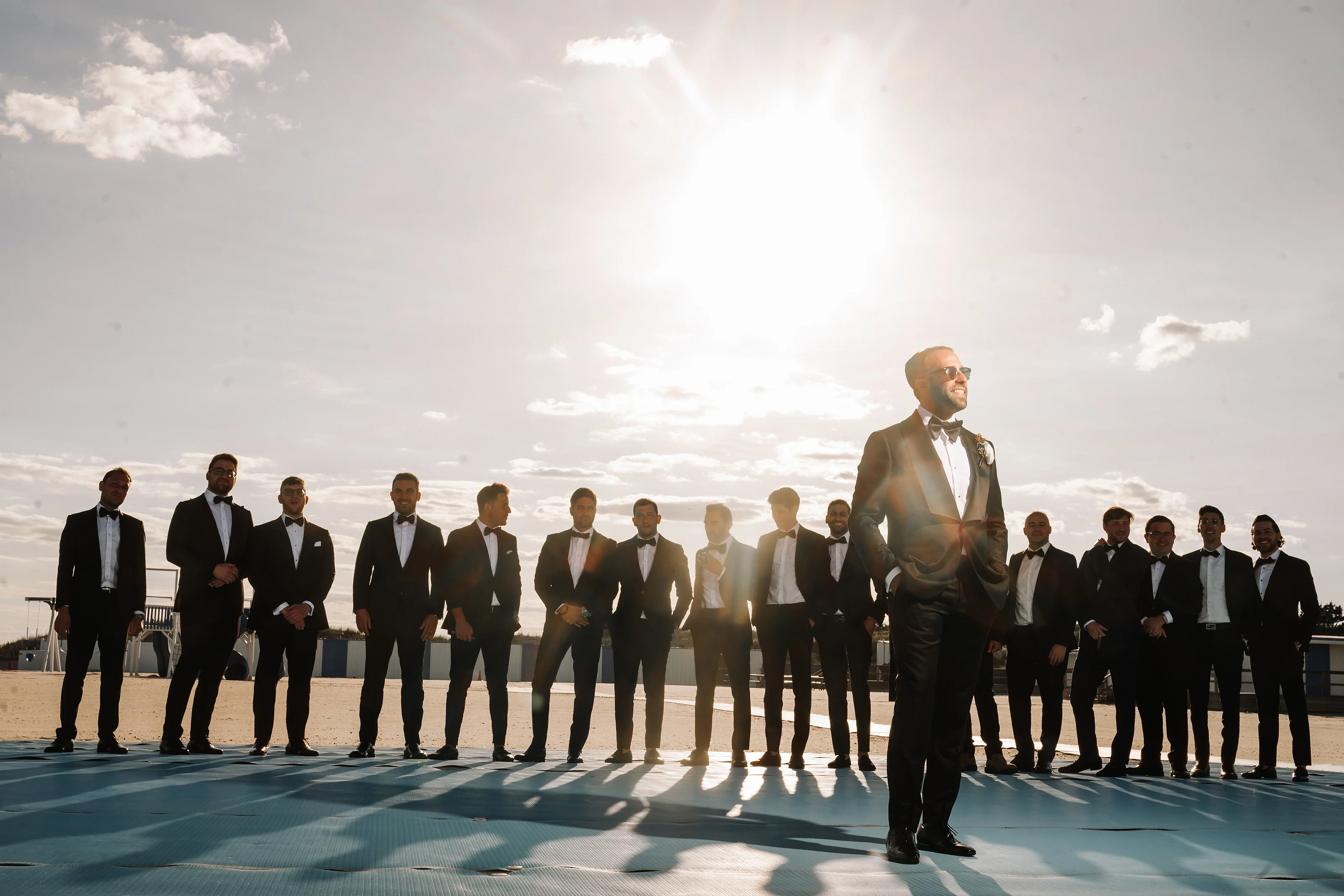 Backlit wedding party group standing in a line along a waterfront at sunset, with the groom in front and bright sky behind.