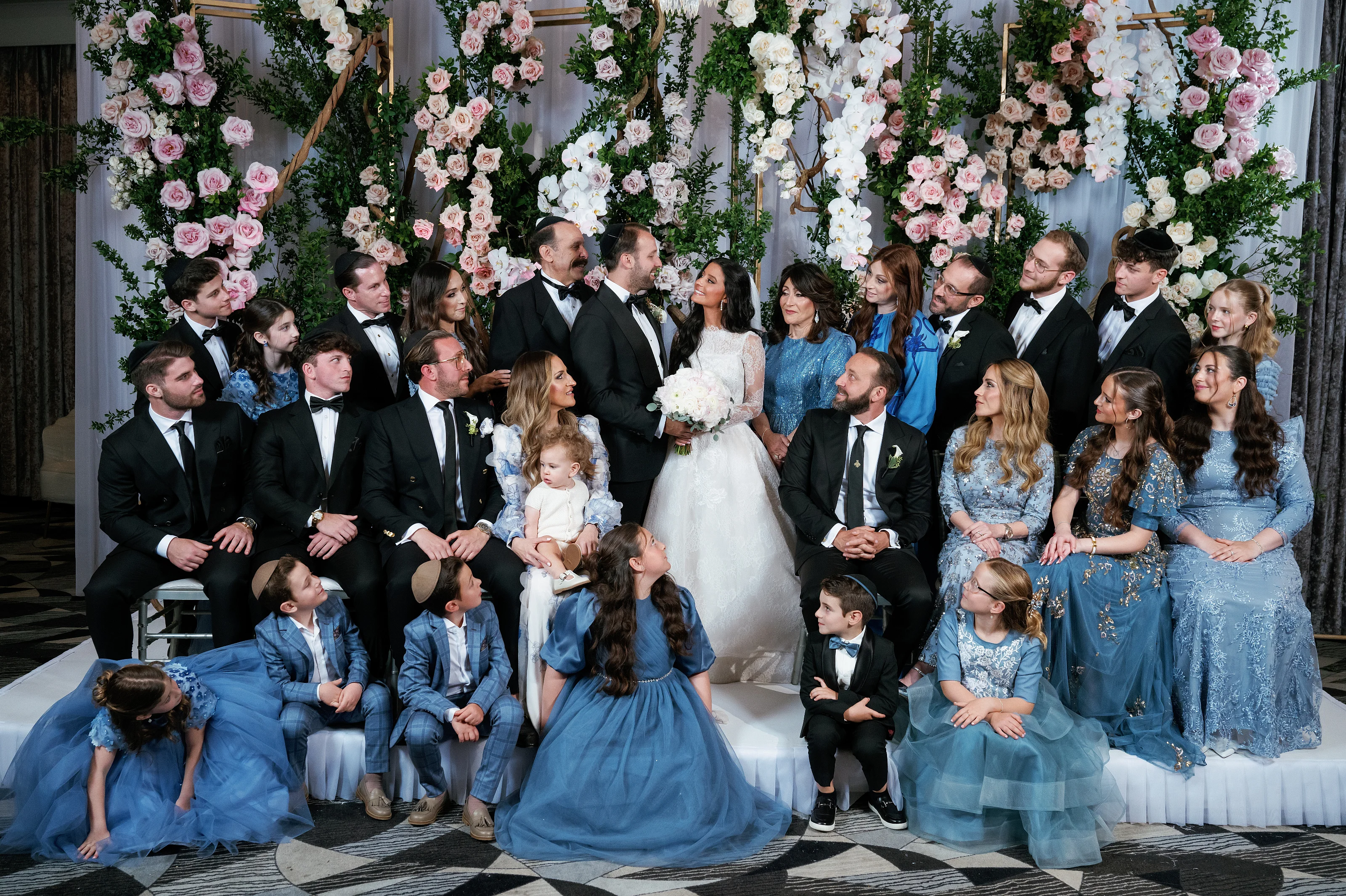 The bride and groom pose with immediate family and children in coordinated blue outfits in front of an ornate floral backdrop at a wedding ceremony.