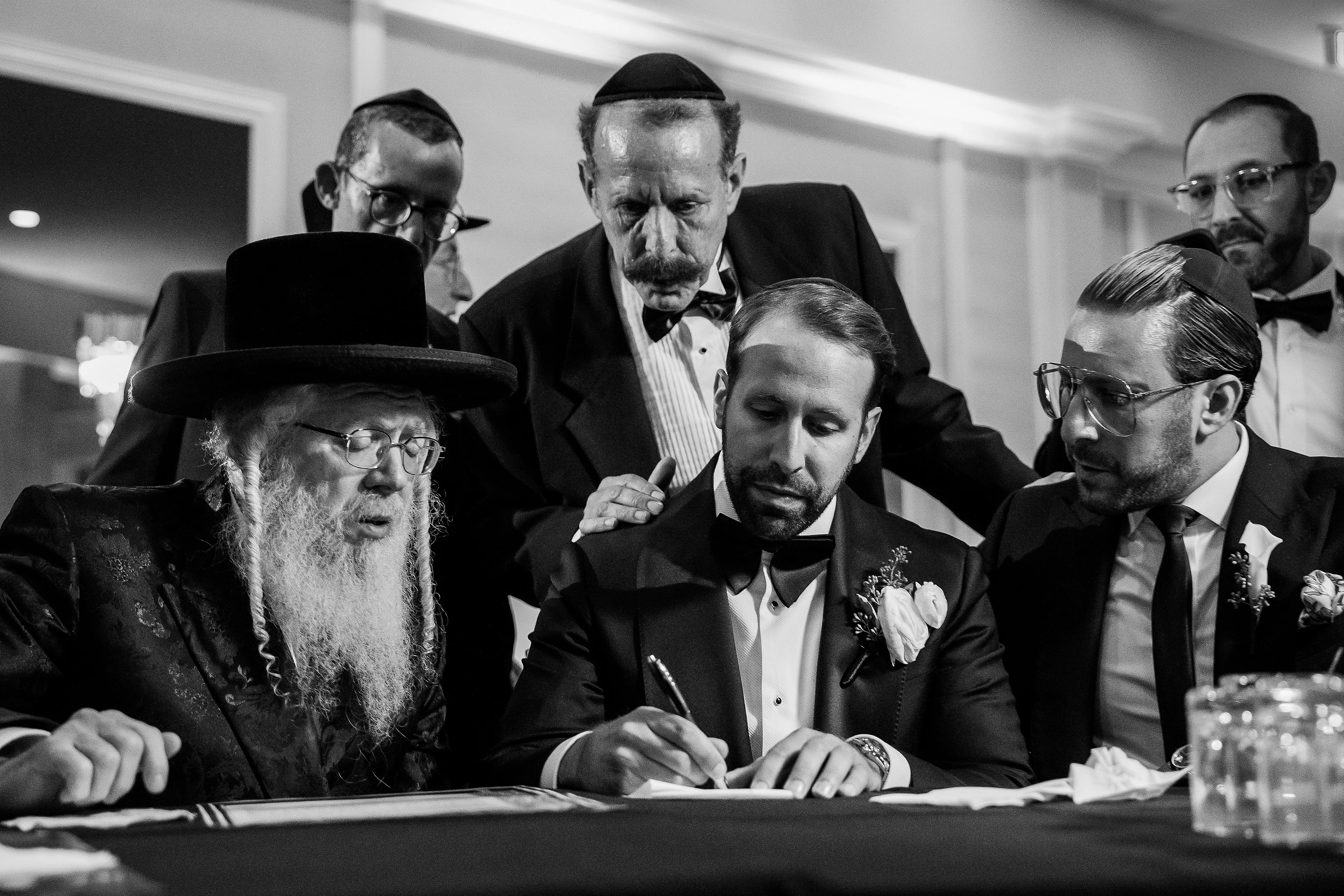 Black-and-white photo of a groom seated at a ritual table during tefillin, writing with close family looking on.