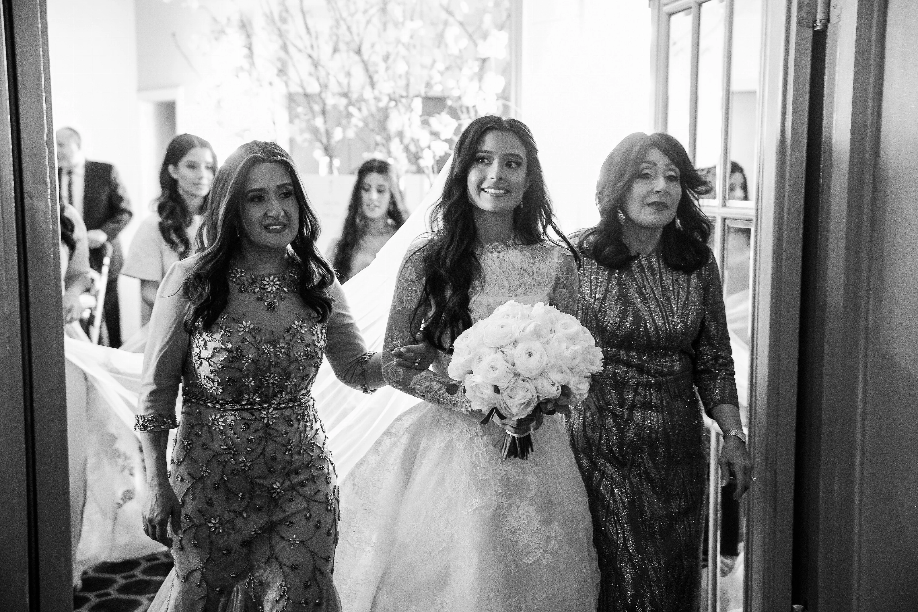 Black-and-white photo of a bride holding a bouquet as she steps forward with family in a bright indoor hallway.