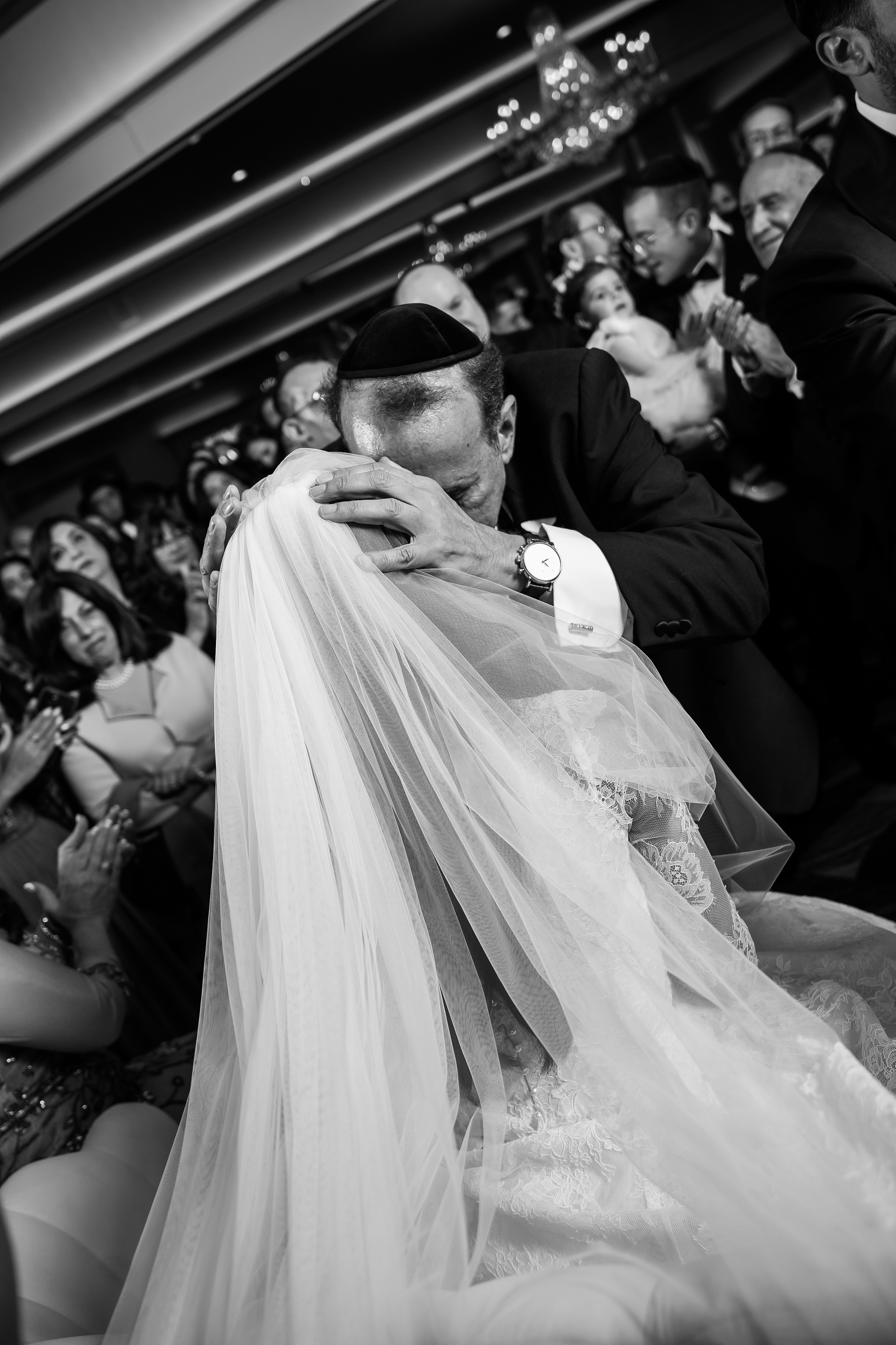Black-and-white photo of a groom embracing with his bride during the badeken, veil flowing as guests watch from behind.