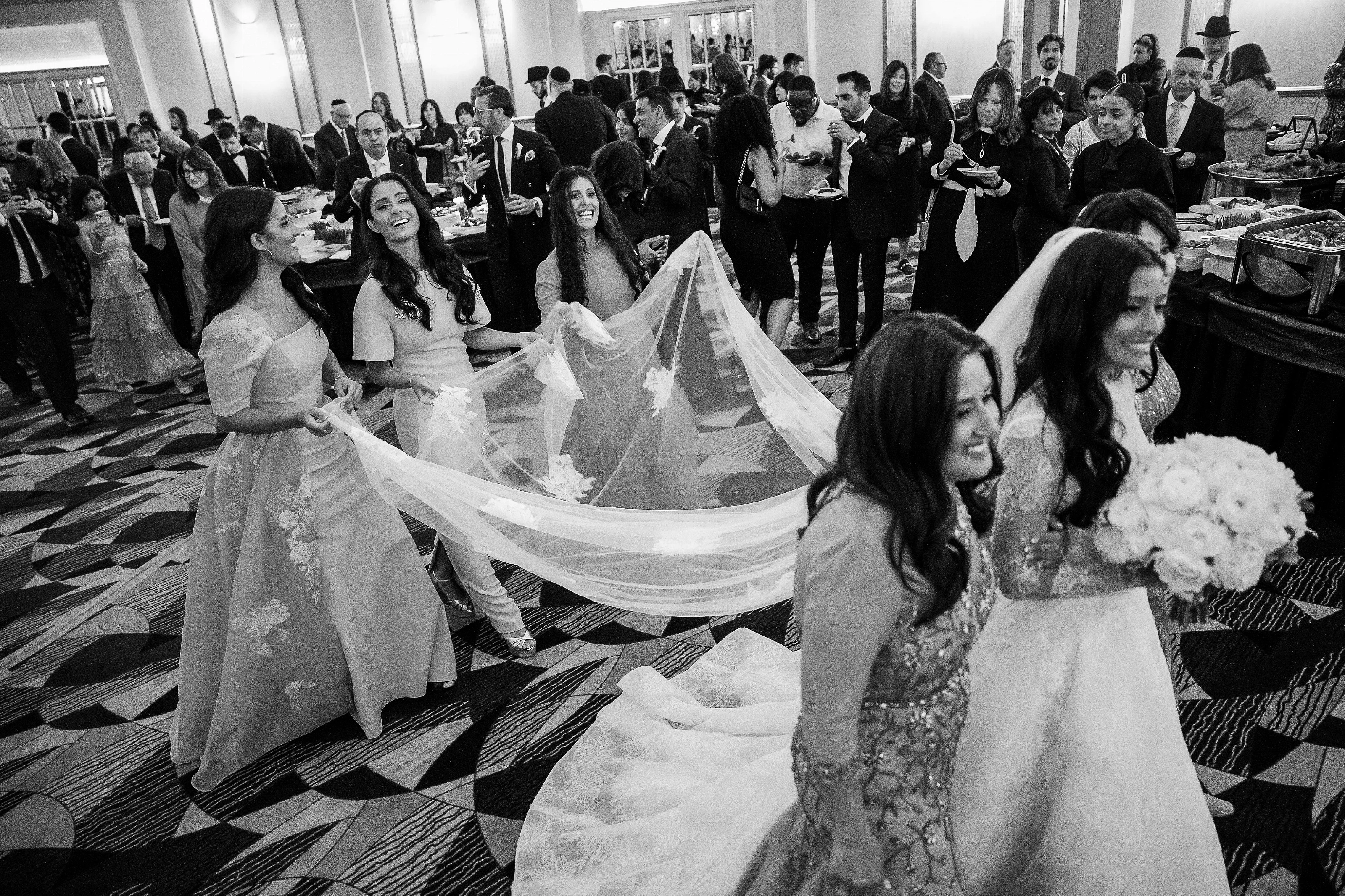 Black-and-white reception photo showing guests smiling during the dance floor celebration, with wedding veil fabric visible in the foreground.
