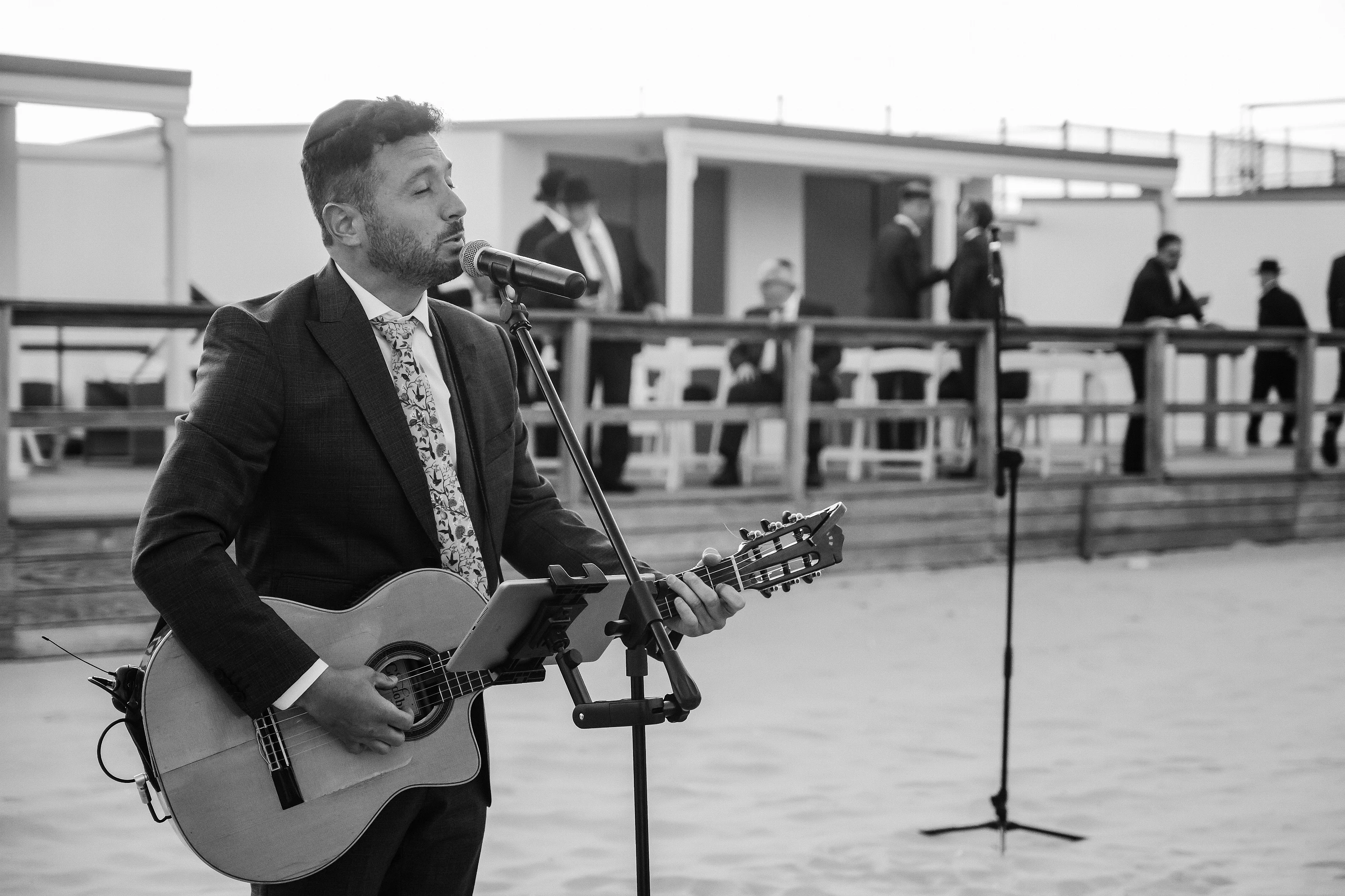 Black-and-white photo of a guitarist performing into a microphone outdoors, with blurred musicians in the background.