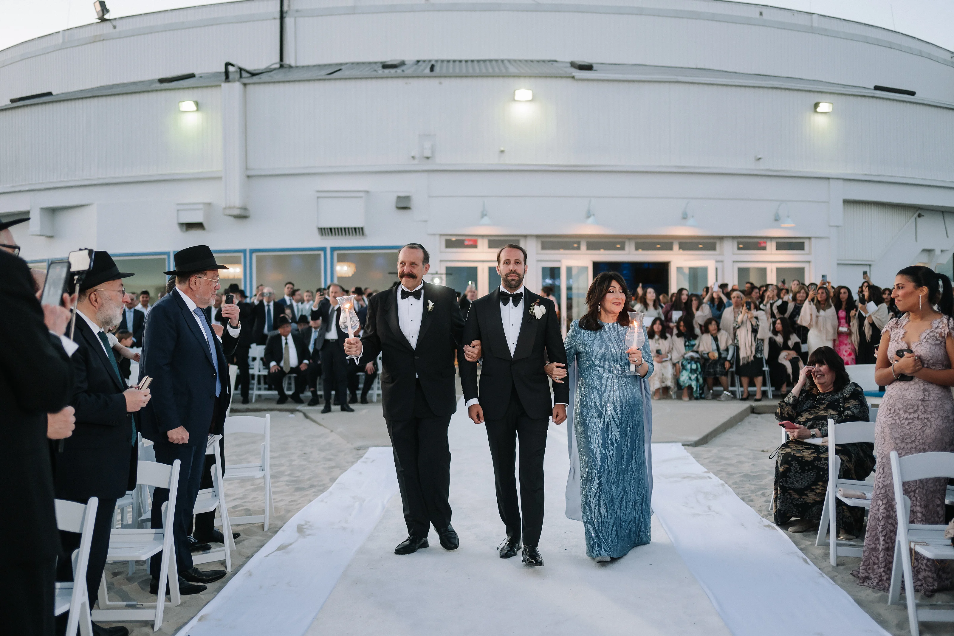 Orthodox Jewish wedding entrance with the couple walking down the aisle toward the camera while guests watch from both sides.