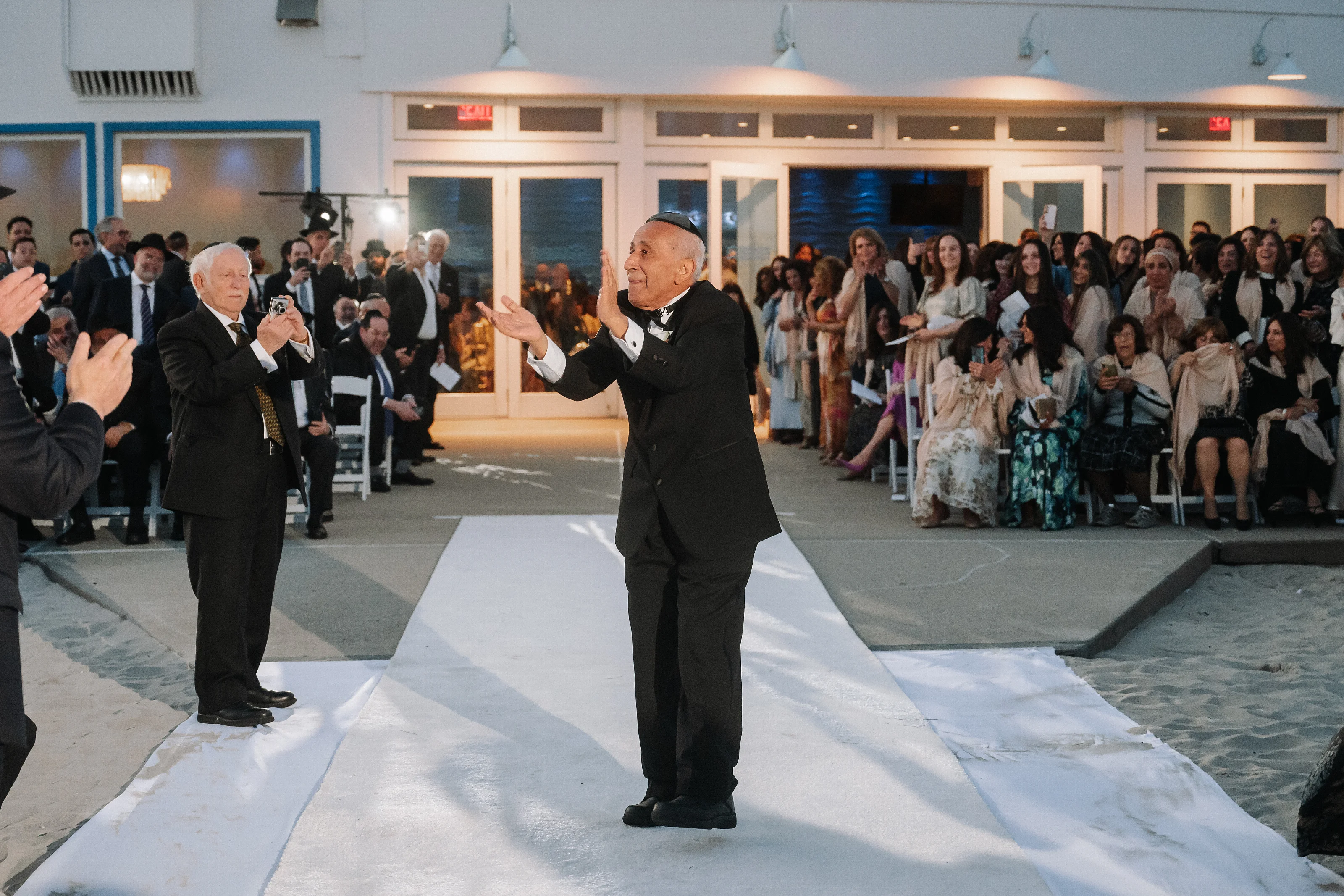 Two men in formal attire gesture on a reception aisle while guests watch from both sides.