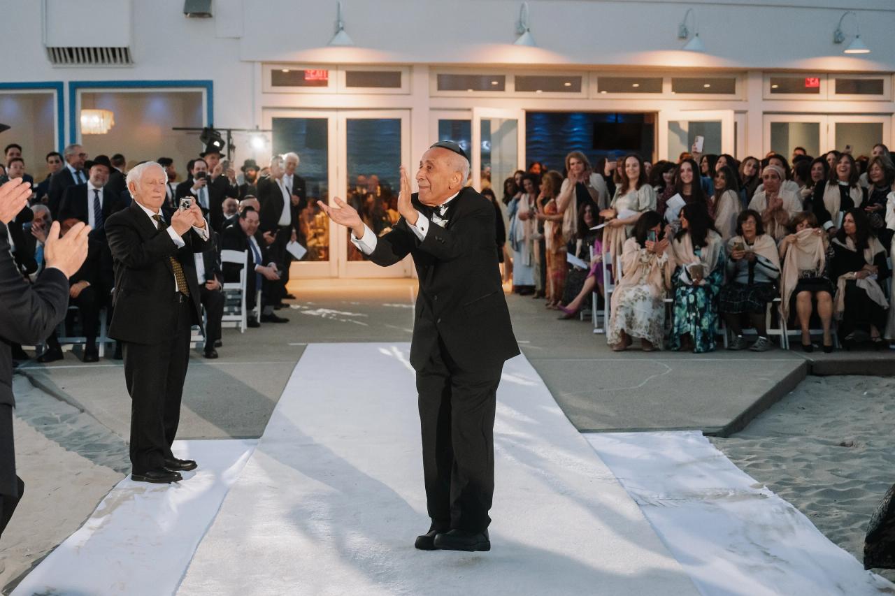 Two men in formal attire gesture on a reception aisle while guests watch from both sides. Wedding photography at Sands, Atlantic Beach, NY.