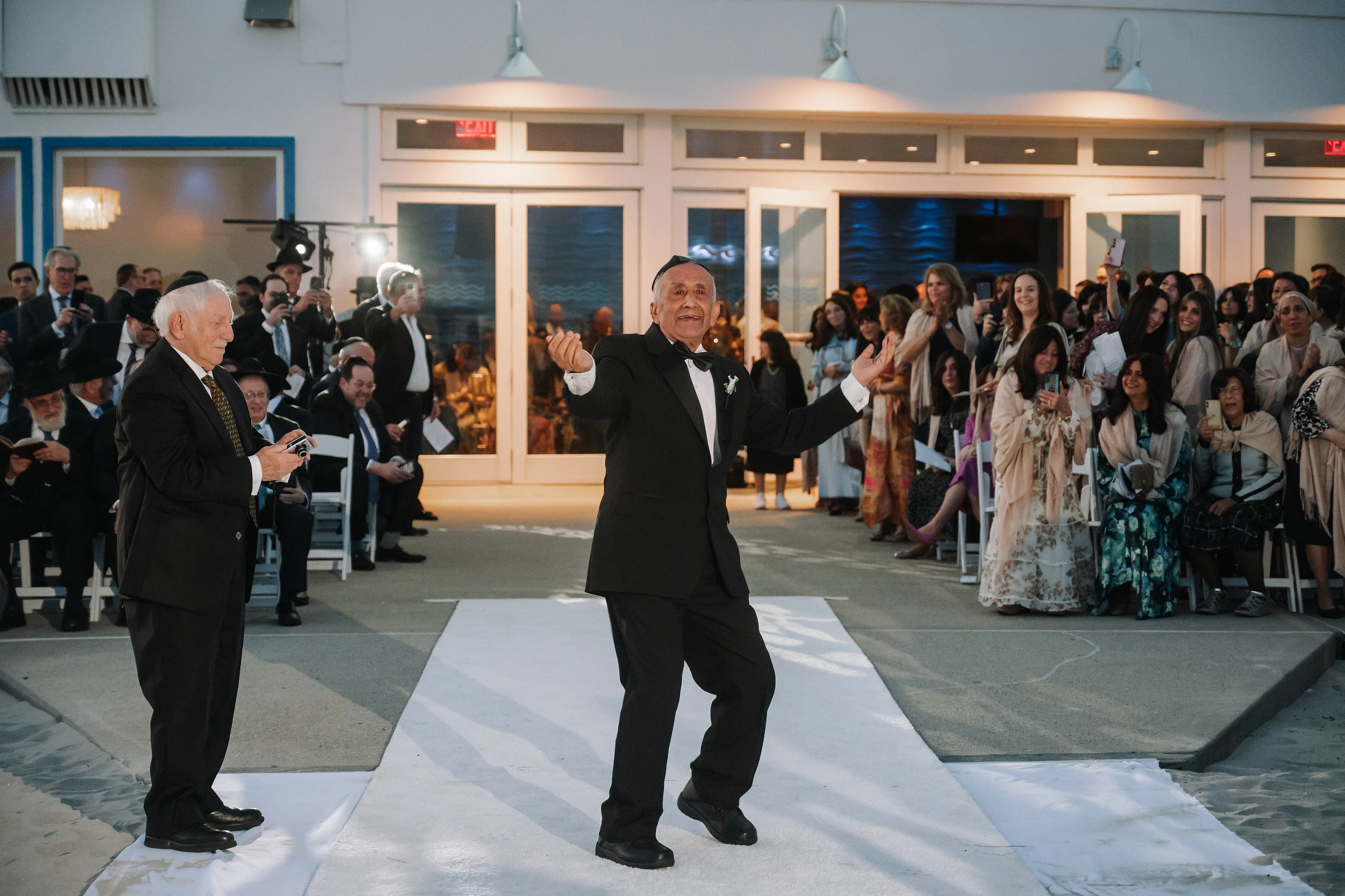 Two men in formalwear dancing on the wedding venue floor as guests gather and clap in the background.