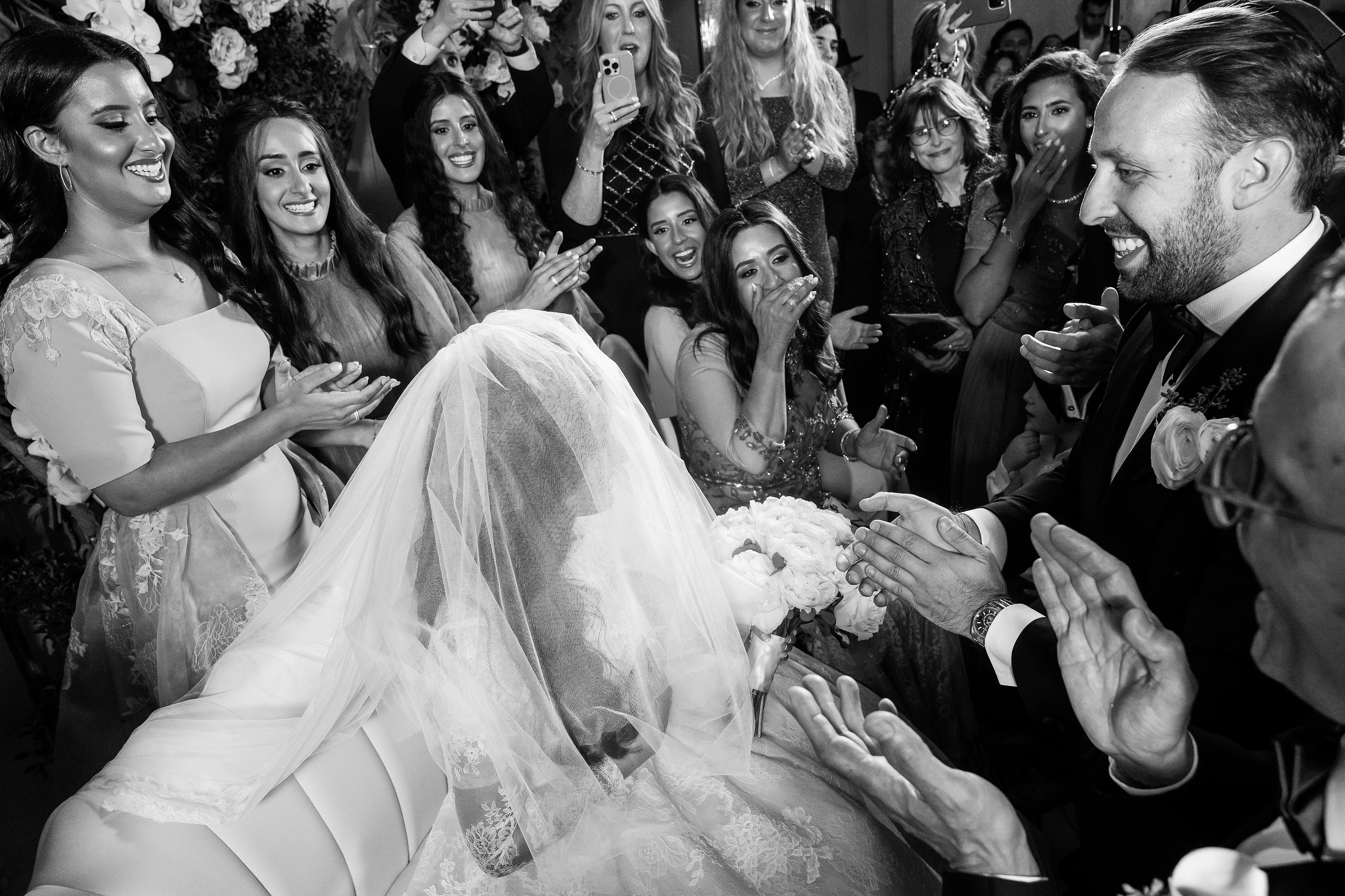 Black-and-white reception moment with bride and groom smiling as the bride leans into a cheering crowd, veil flowing toward the camera.