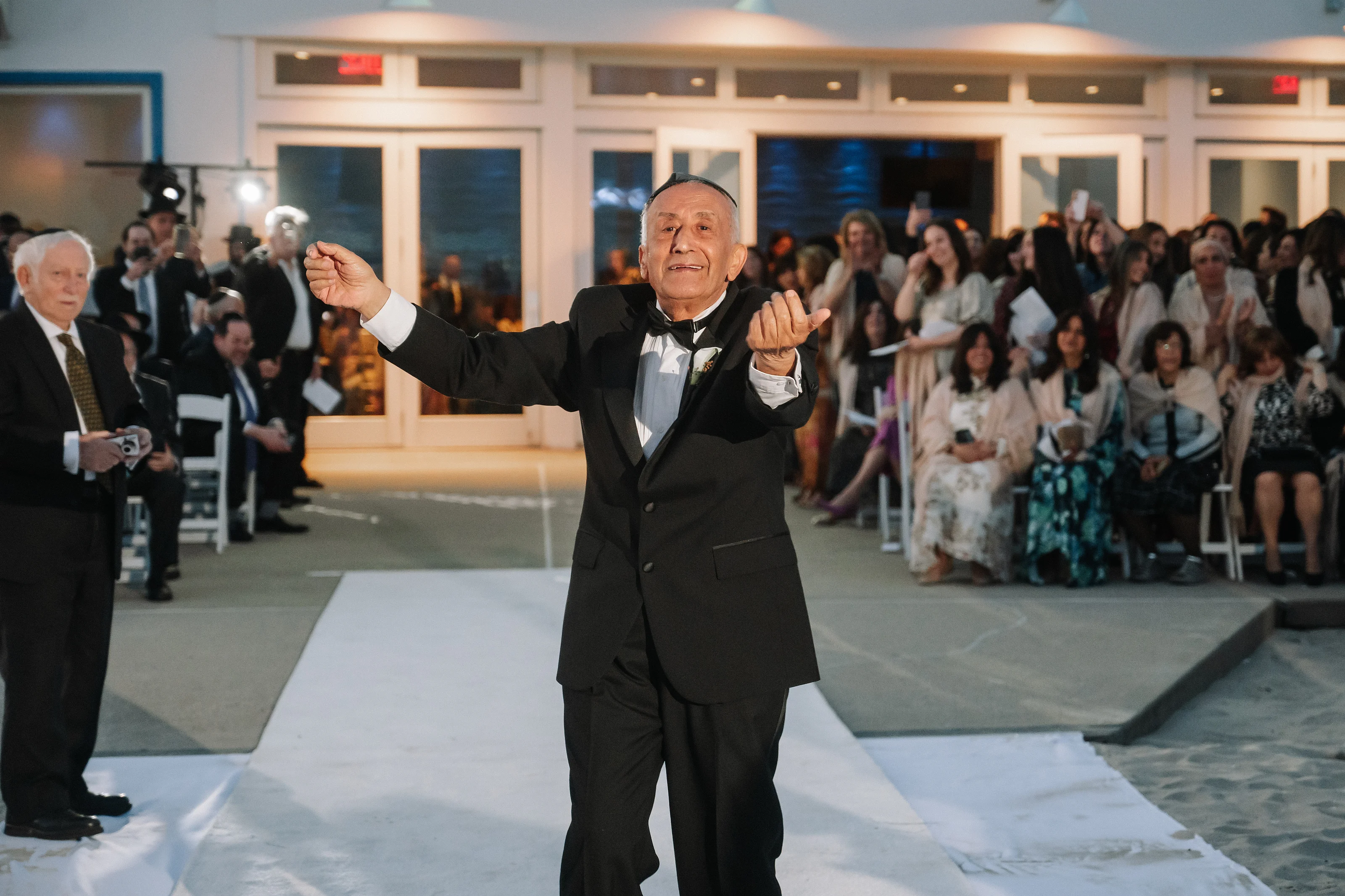 Smiling man in a tuxedo making a celebratory hand gesture on a ballroom dance floor while seated guests watch in the background.